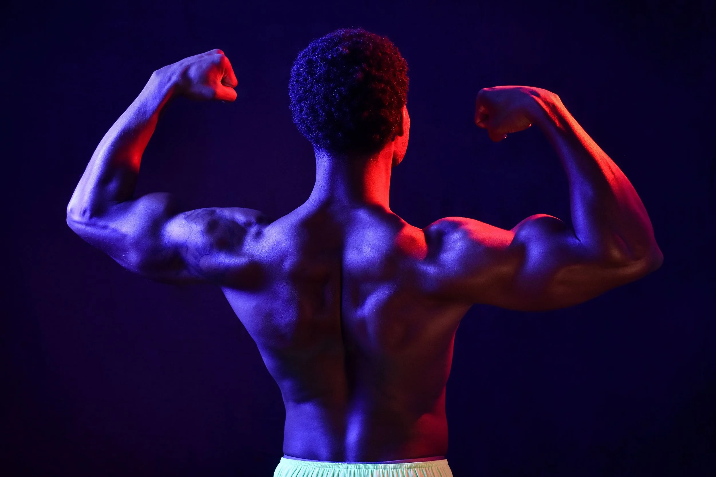 A male personal trainer poses in front of a black backdrop, facing away from the camera. He flexes his biceps and deltoid muscles, which are illuminated and defined by the red and blue lights on both sides.