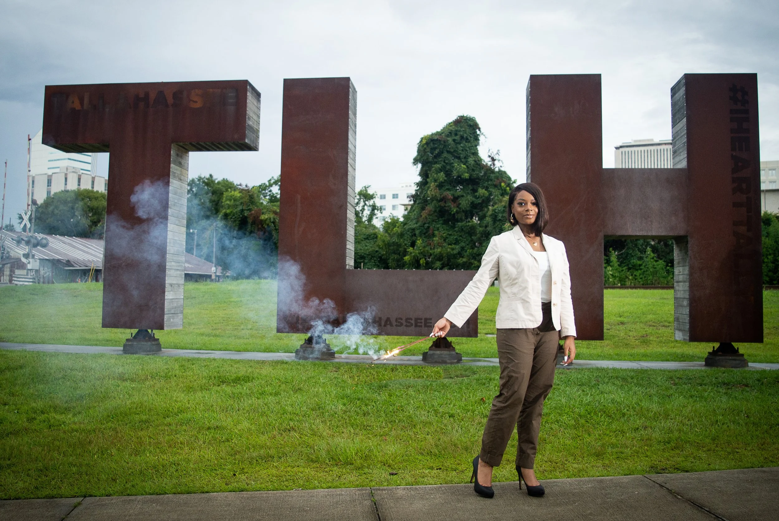A young Black woman wearing a cream-colored blazer and chocolate brown slacks, stands in front of large copper and wooden "TLH"  letters, holding 2 sparklers.