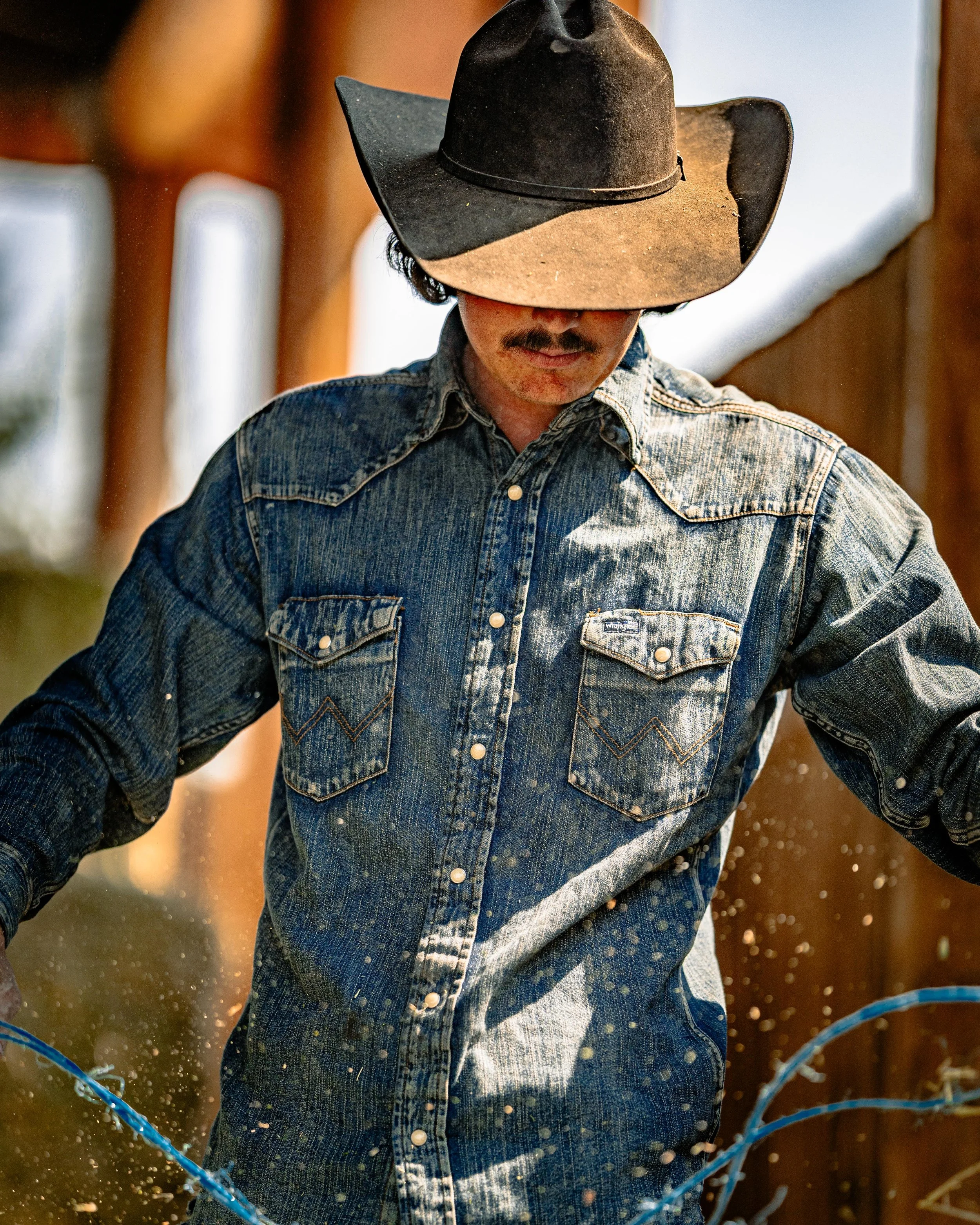 A man wearing a black cowboy hat, a denim shirt, and a mustache, looking down while holding a blue rope outdoors.