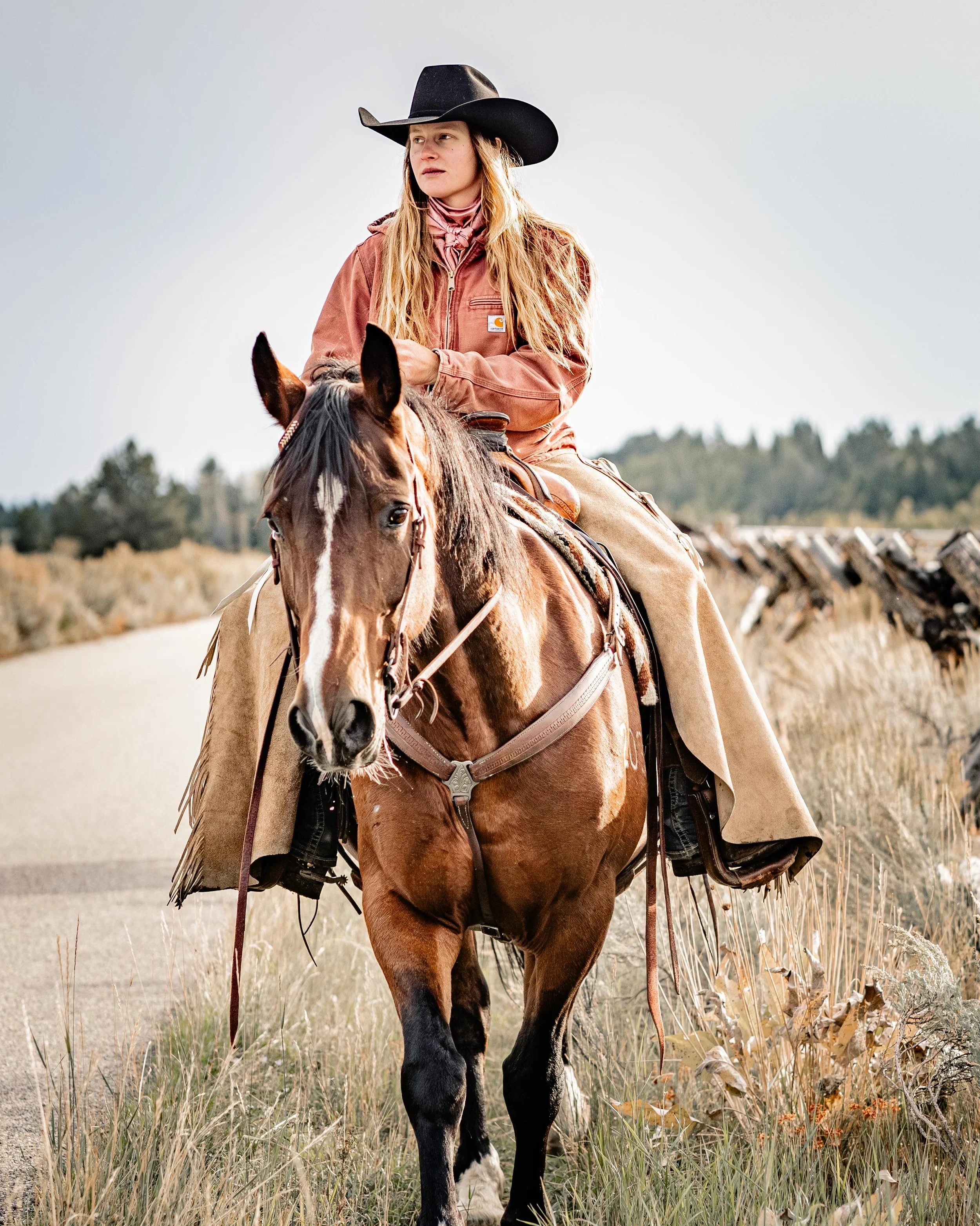 A woman wearing a black cowboy hat and a brown jacket riding a brown and white horse on a rural dirt path.