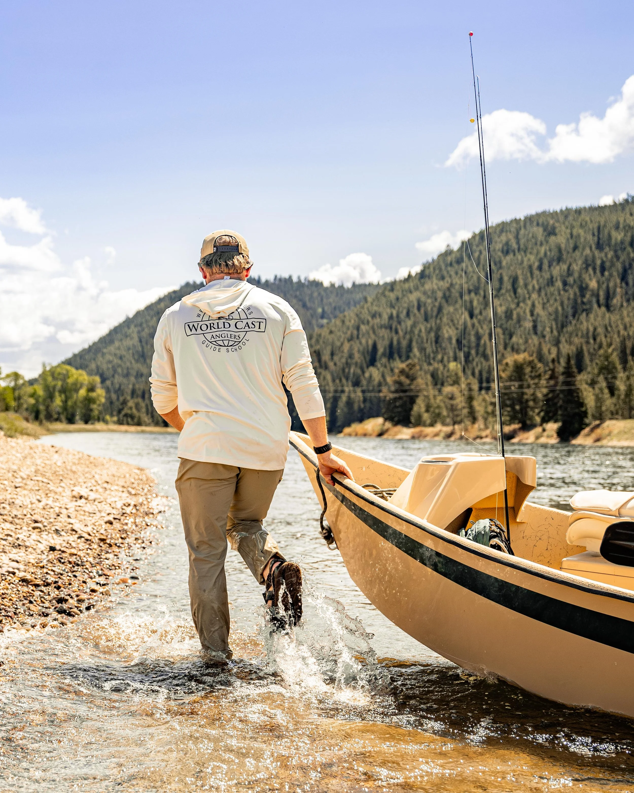 A person with a beige cap and a white hoodie pulling a small yellow boat out of the water onto a rocky shoreline, with a river, green hills, and a blue sky in the background.