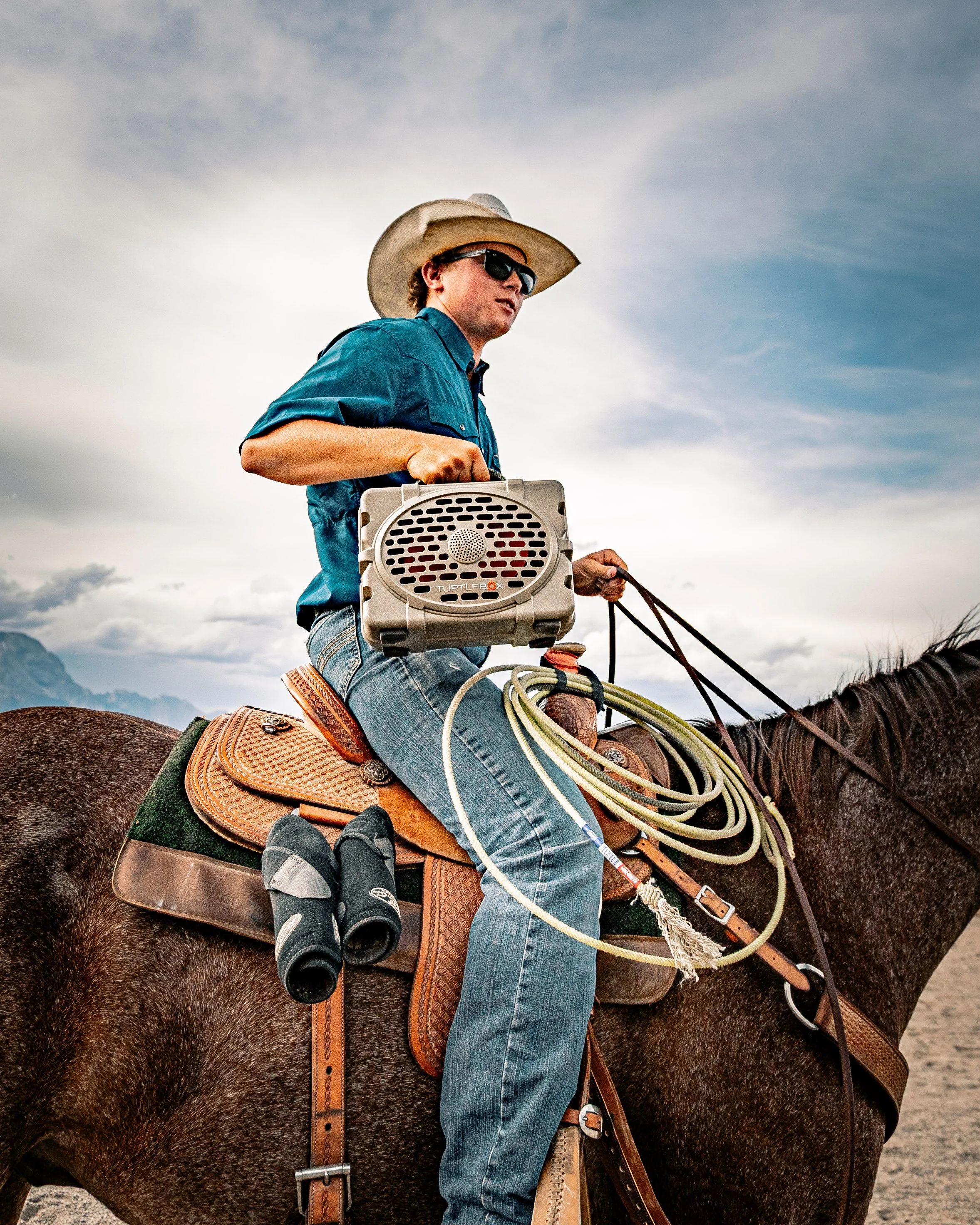 A man wearing a cowboy hat, sunglasses, and a blue shirt rides a horse. He holds a portable speaker in one hand and reins in the other, with a lasso and binoculars attached to his saddle. The sky is partly cloudy.