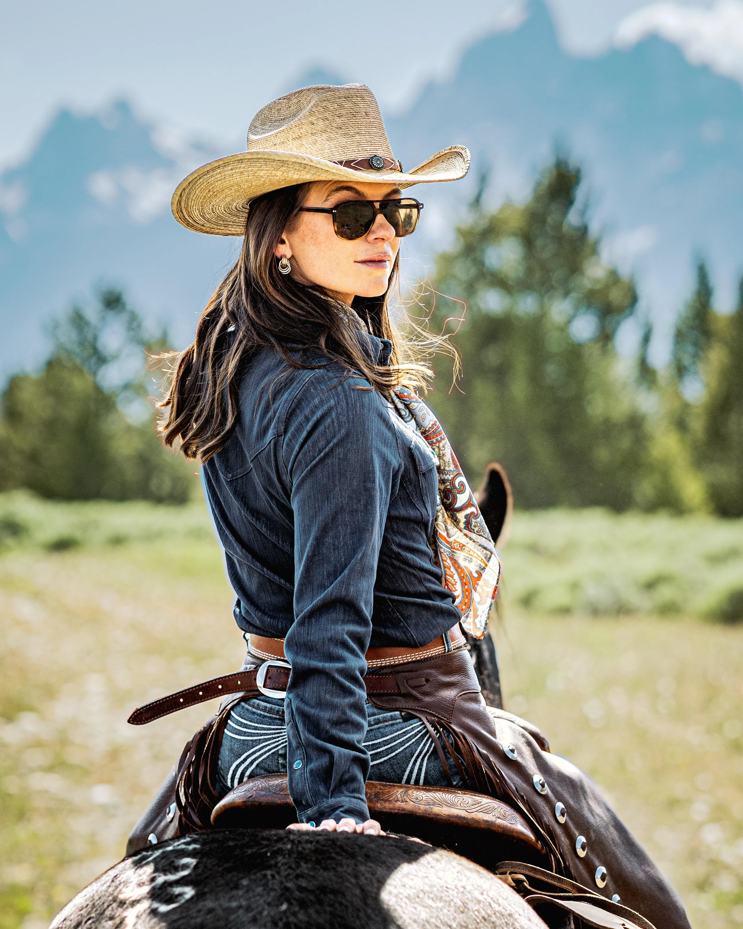 A woman wearing sunglasses, a cowboy hat, a denim shirt, and a scarf, riding a horse outdoors with mountainous terrain and trees in the background.