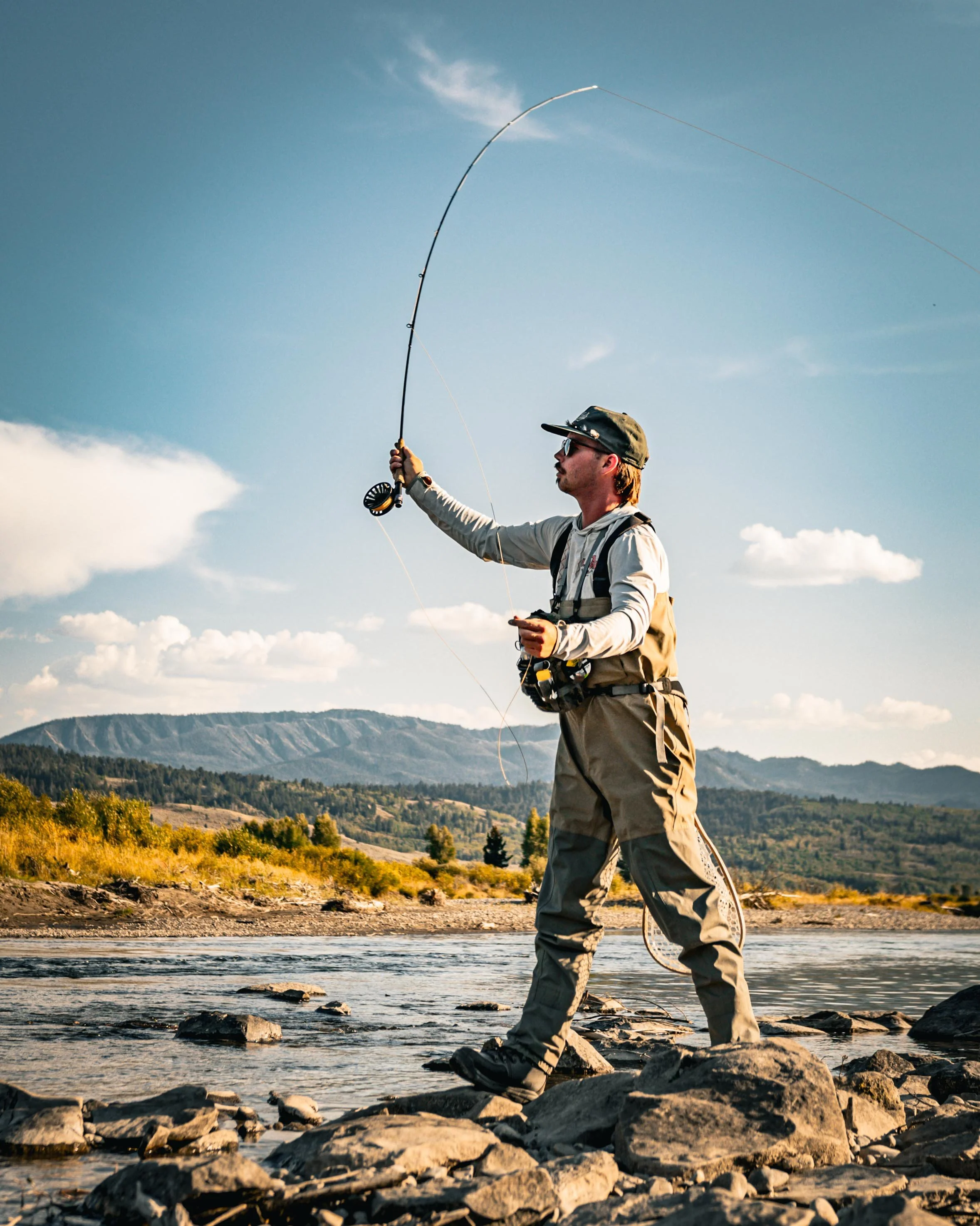 Man fishing in a river with mountains in the background during daytime.