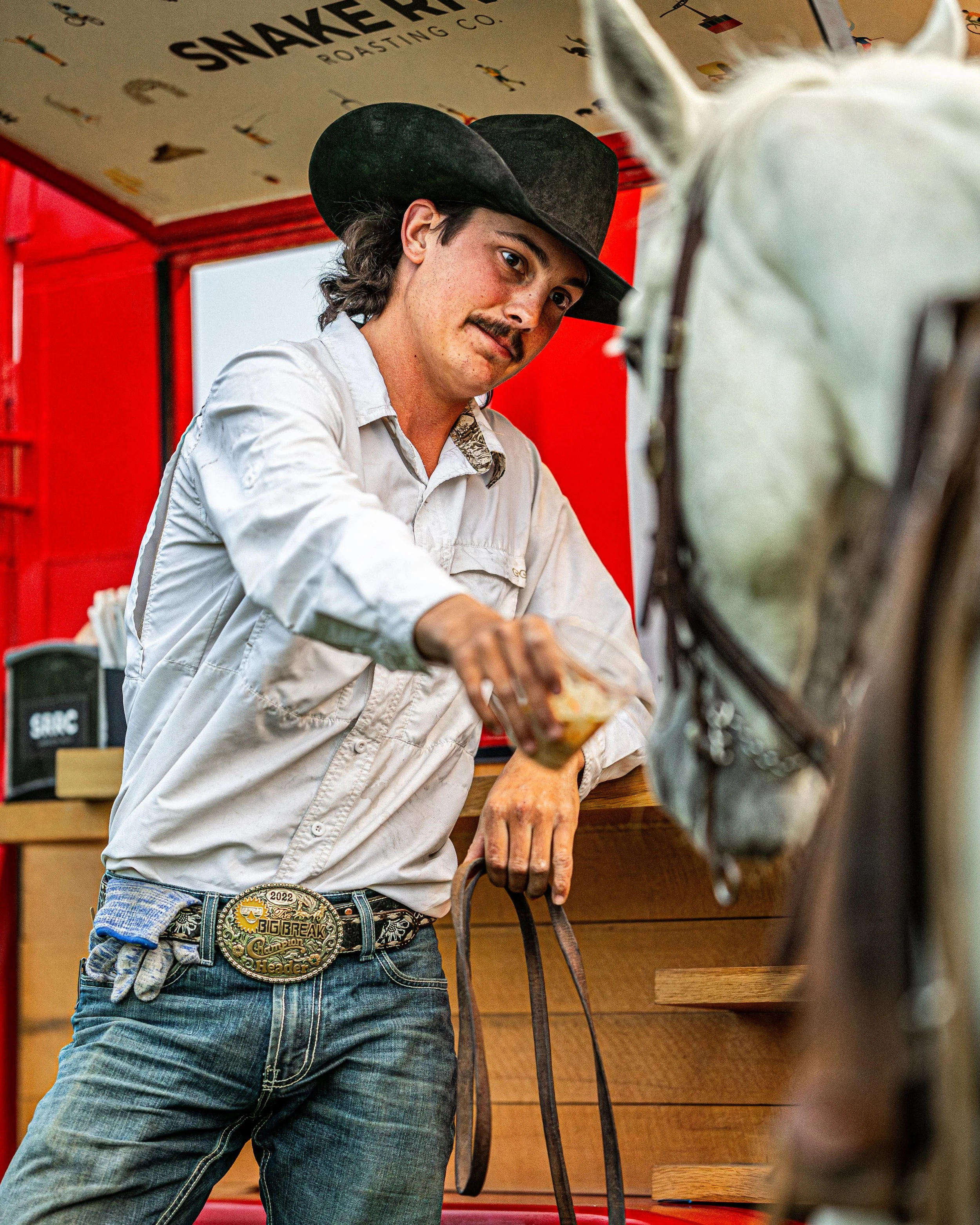 A man with a mustache and long hair wearing a black cowboy hat, a white button-up shirt, and jeans with a large belt buckle, is feeding a white horse in a red-themed setting.