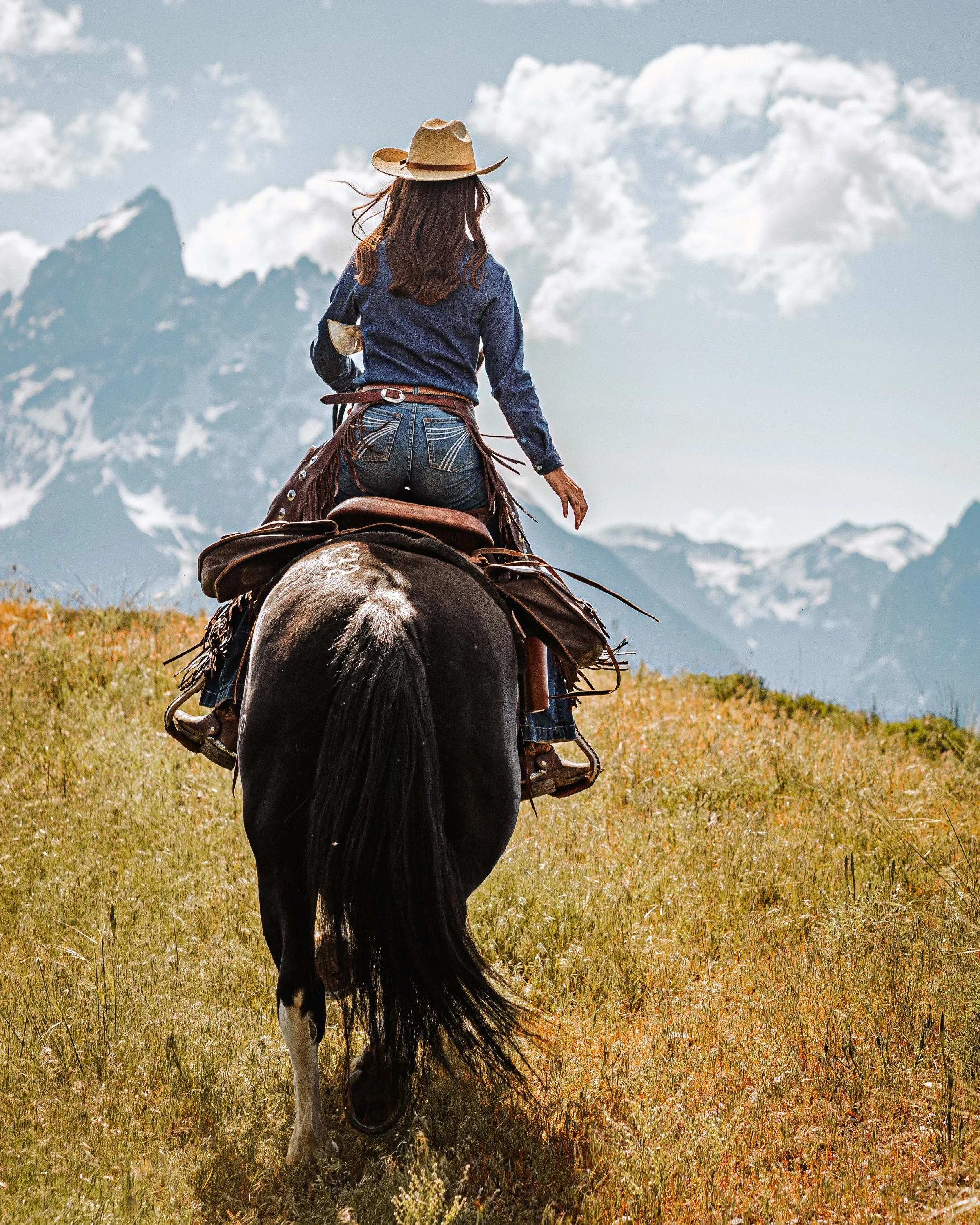 A woman riding a horse in a grassy field with snowy mountains and cloudy sky in the background.