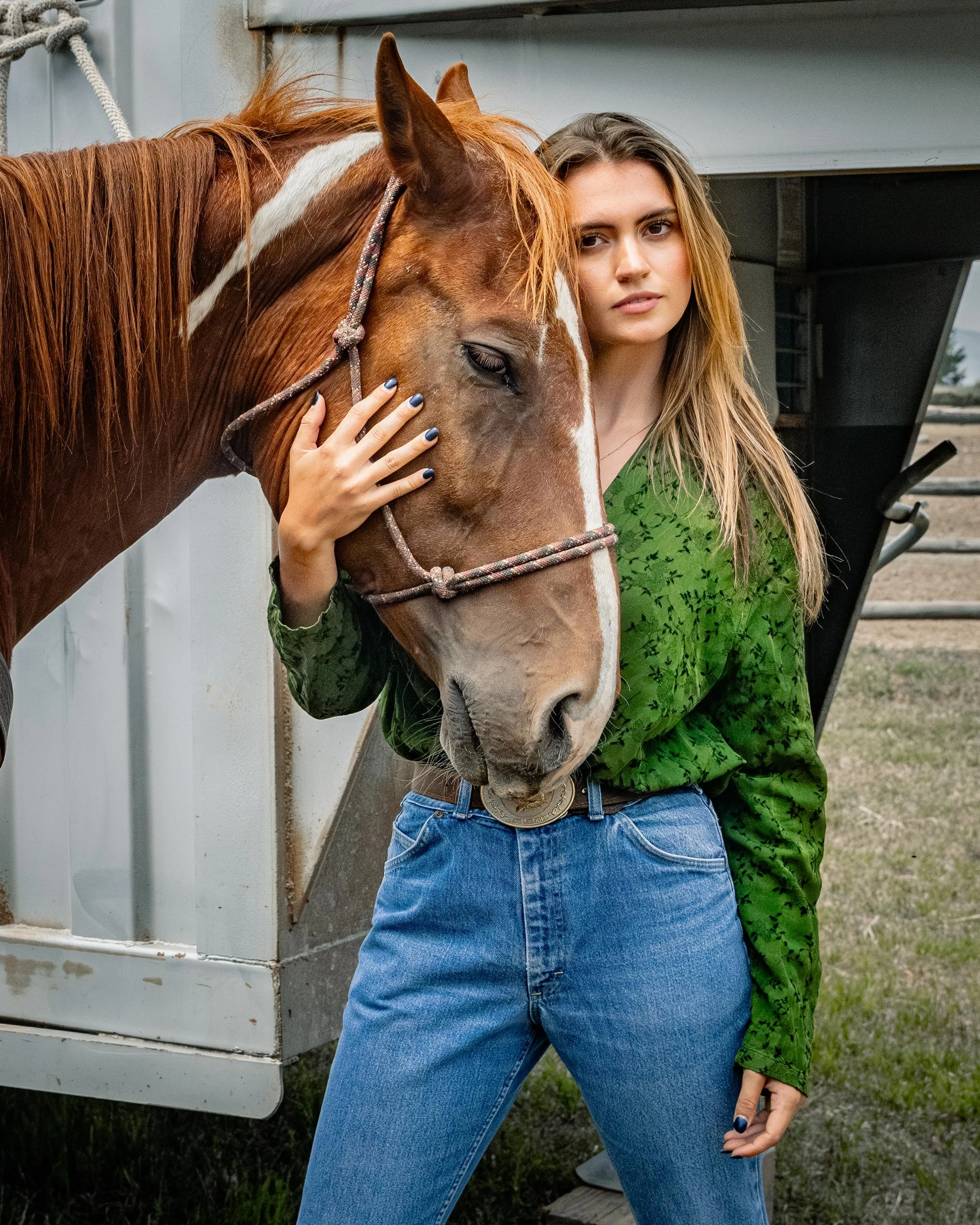 A young woman in a green patterned blouse and blue jeans standing outdoors, gently holding a chestnut horse's face with her hand, with both appearing calm and close.