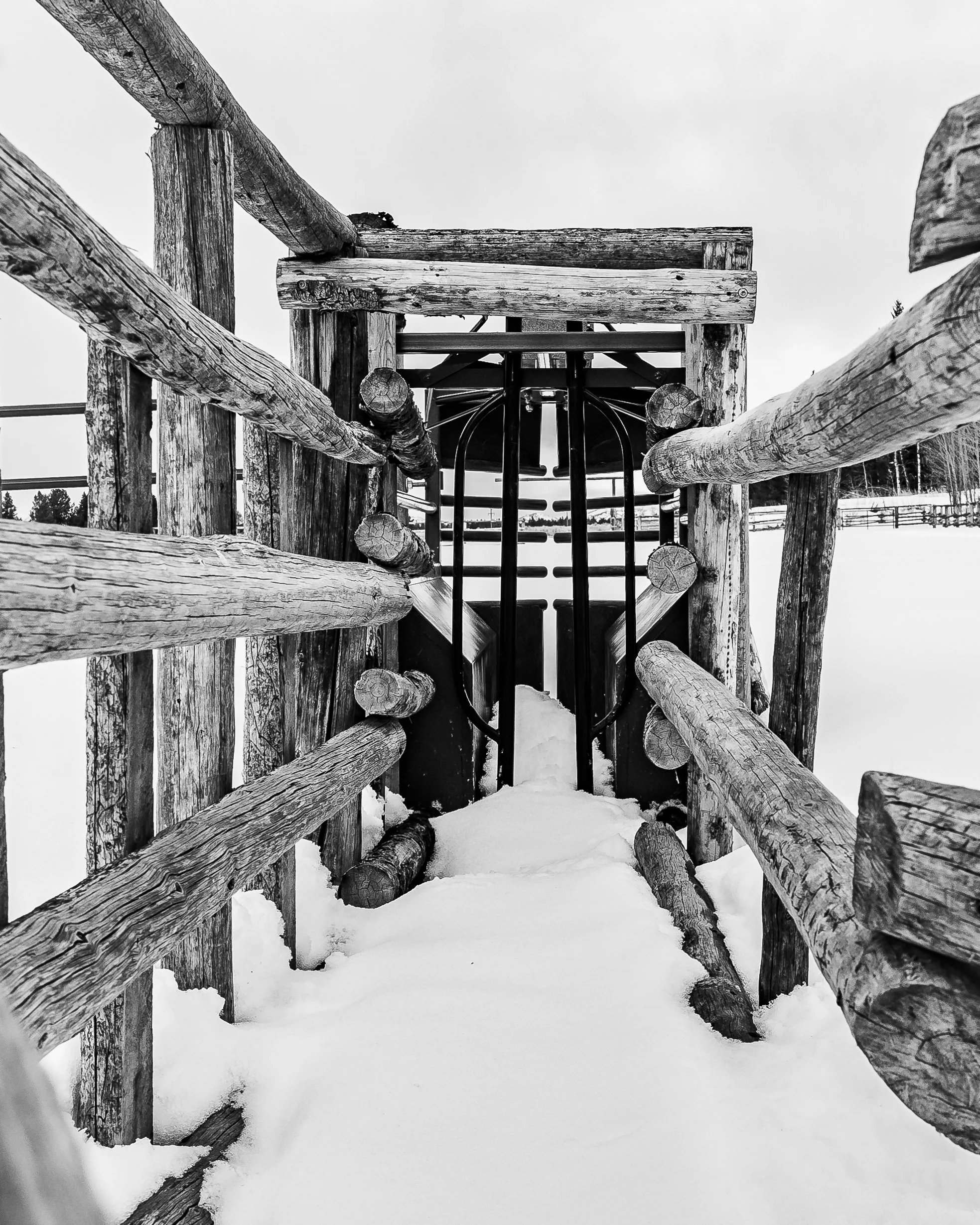 View of a snowy wooden playground structure from the top, showing a slide at the bottom, surrounded by wooden railings and a snow-covered ground.