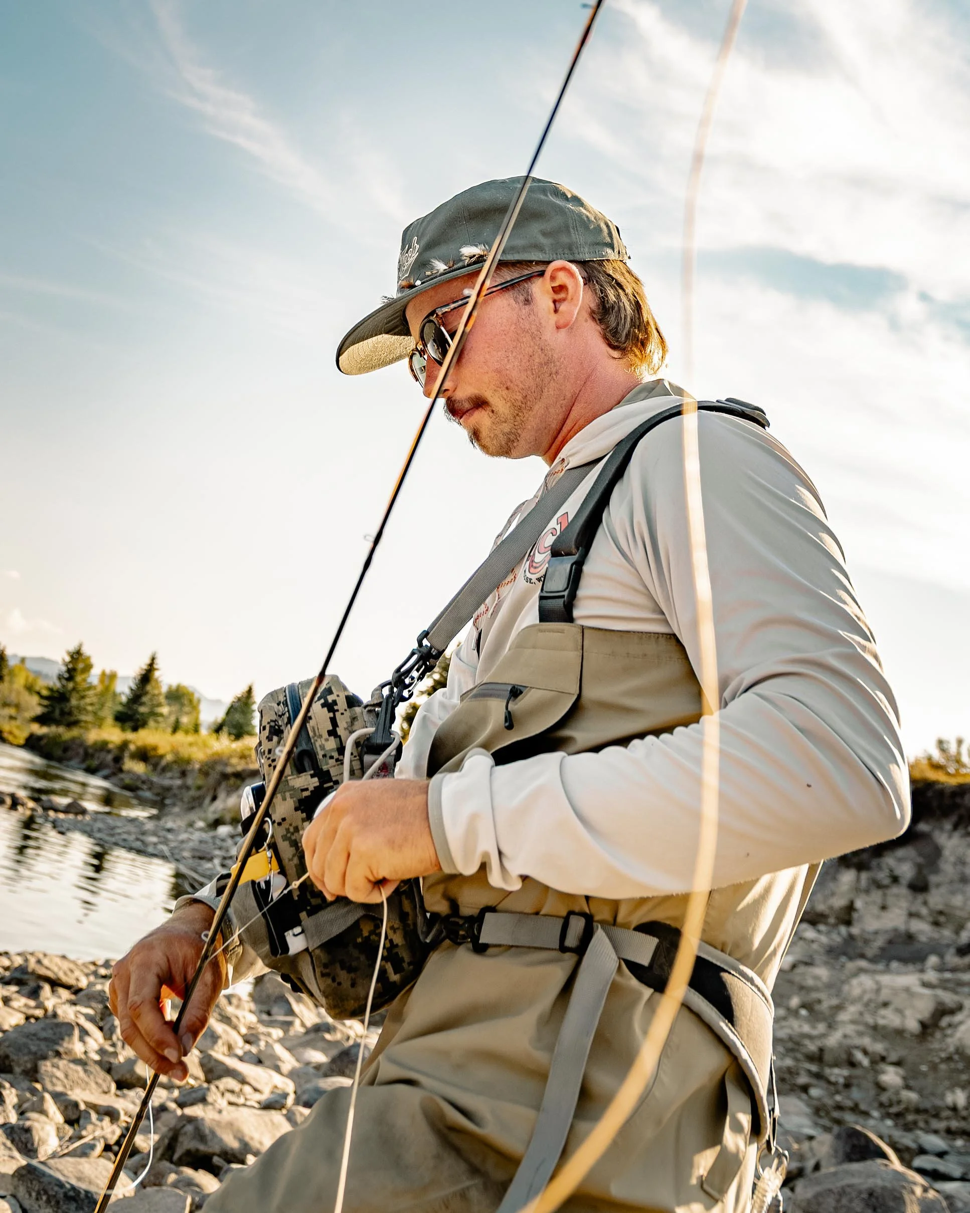 Man fishing by a rocky riverbank during sunset, wearing a hat, sunglasses, and fishing gear.