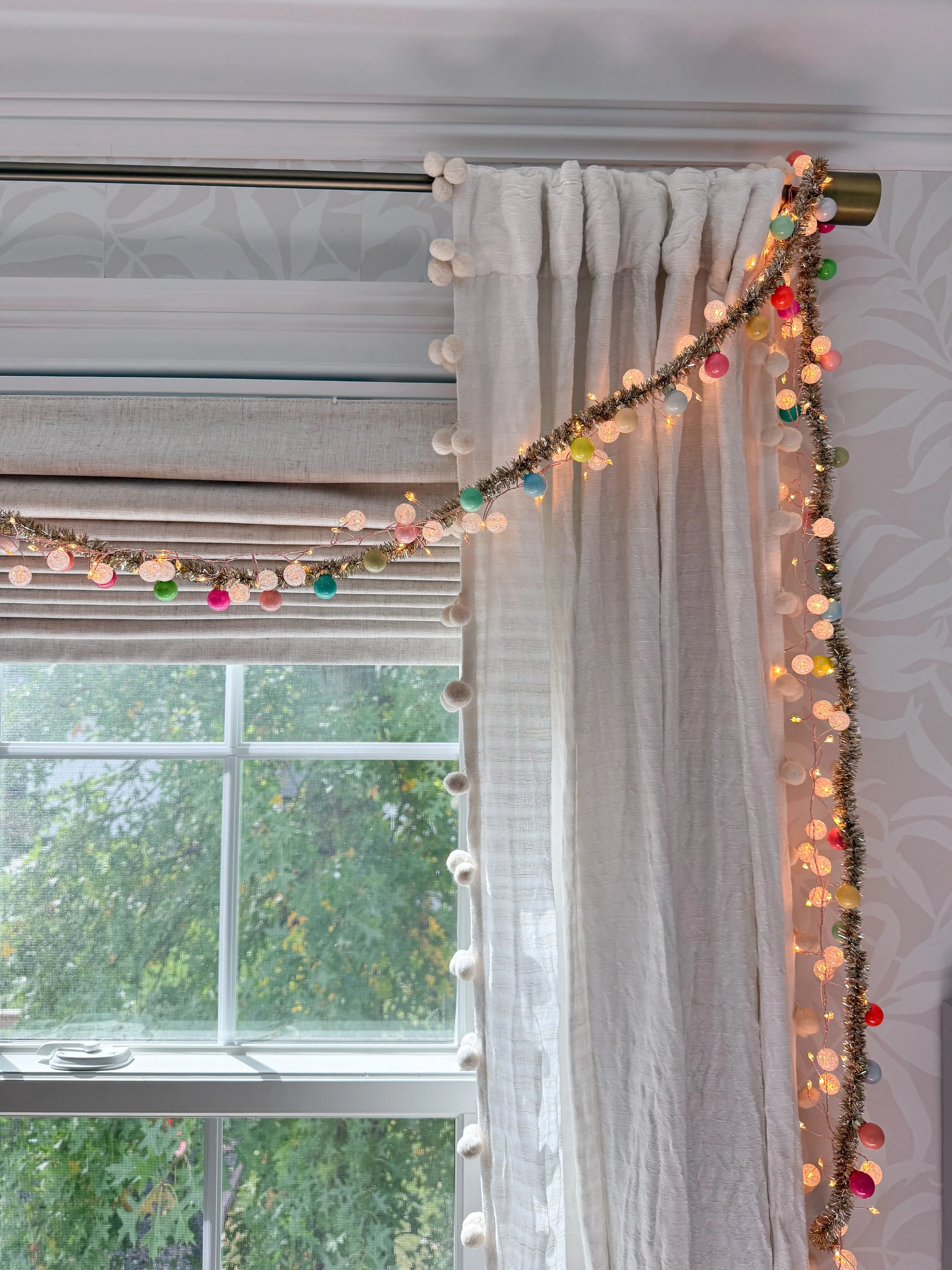 Anthropologie holiday decor featuring colorful multicolored Christmas lights and gold tinsel garland draped across cream pom pom curtains beside a large bedroom window.