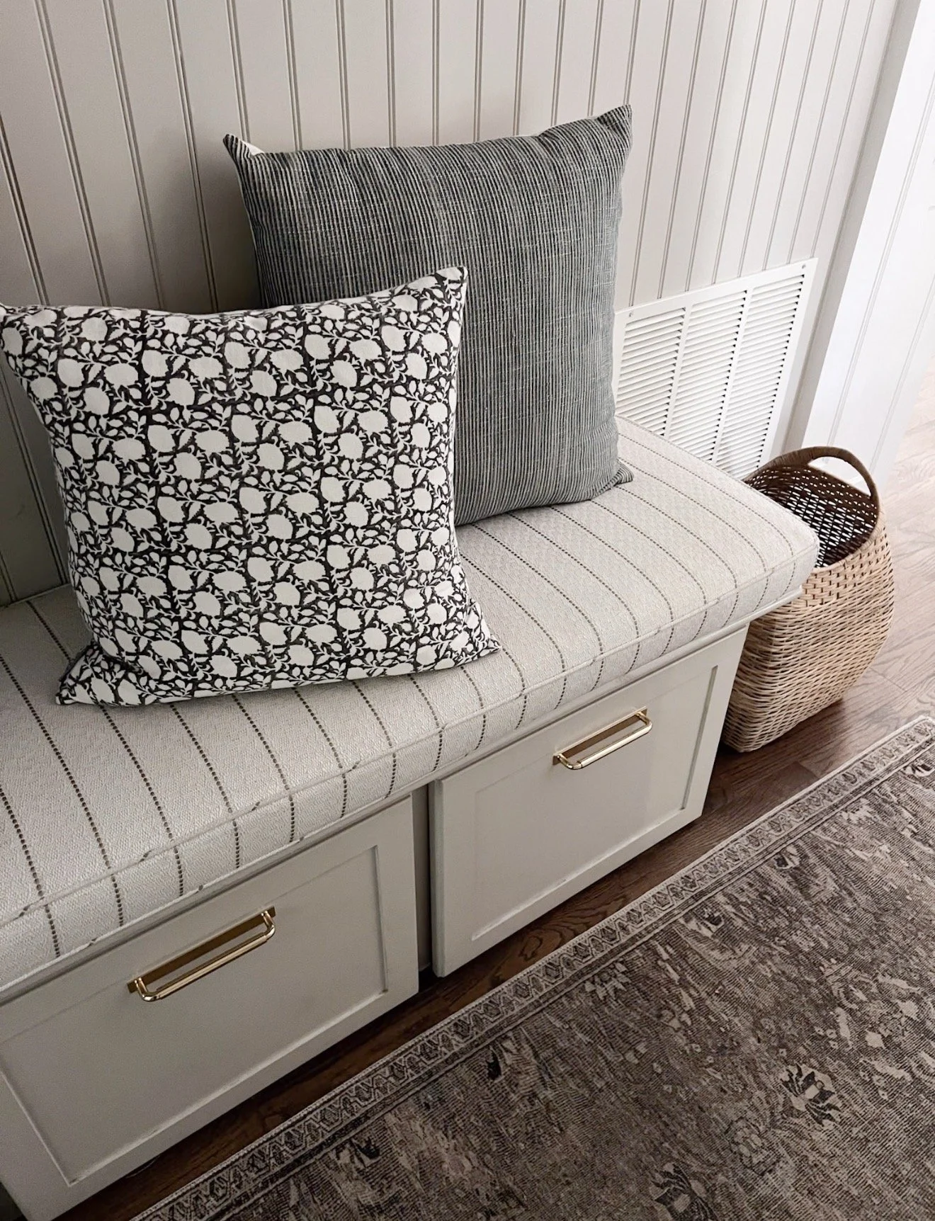 Close-up of a mudroom bench with striped upholstery, patterned pillows, woven basket, and a neutral Loloi runner along the floor.