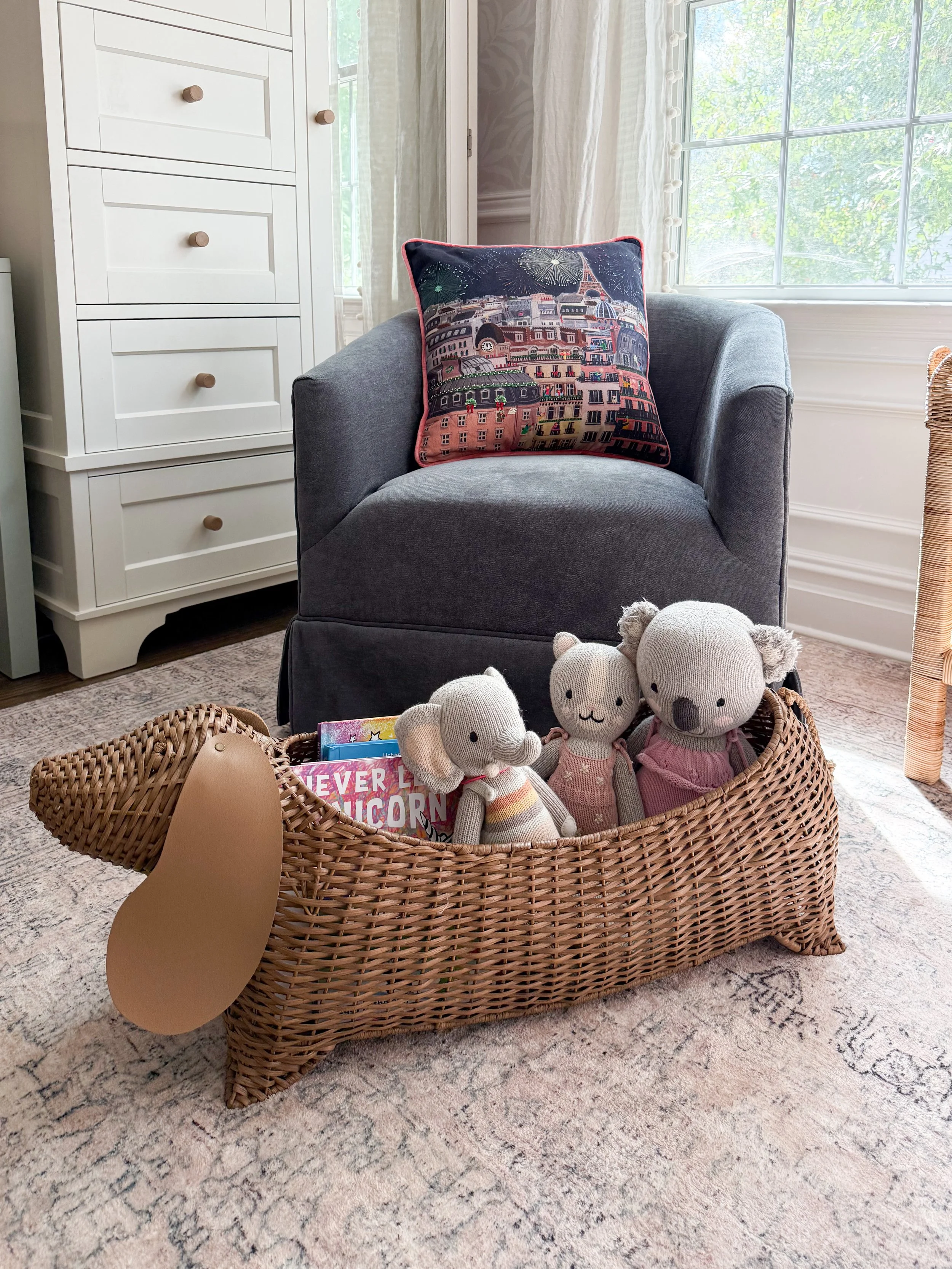 Anthropologie holiday decor featuring a gray accent chair with a colorful Parisian Christmas pillow beside a woven rattan basket filled with plush animal toys and children's books.