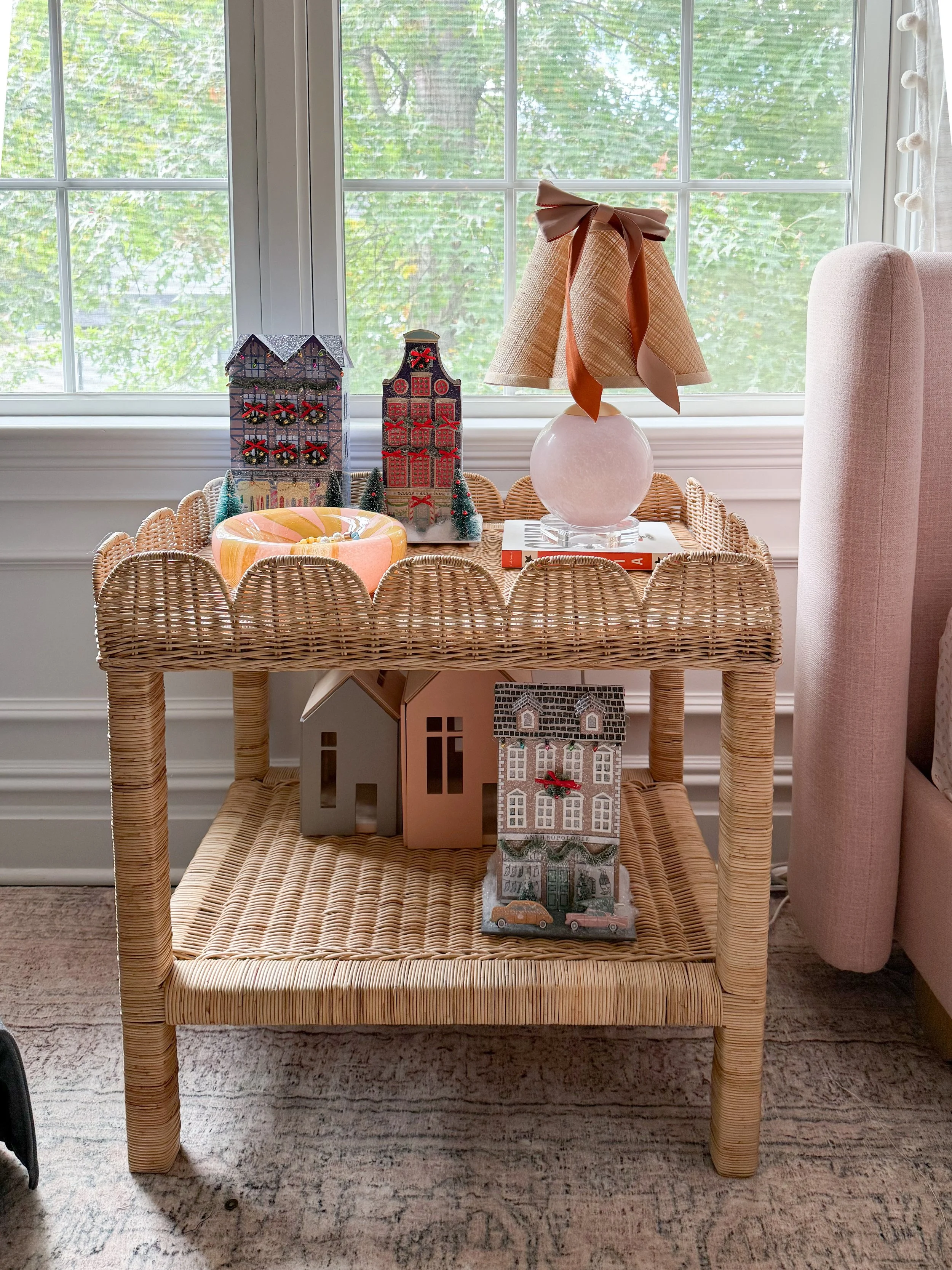 Anthropologie holiday decor featuring miniature Christmas village houses, a pink glass table lamp with a bow, and a colorful decorative dish styled on a woven rattan side table.