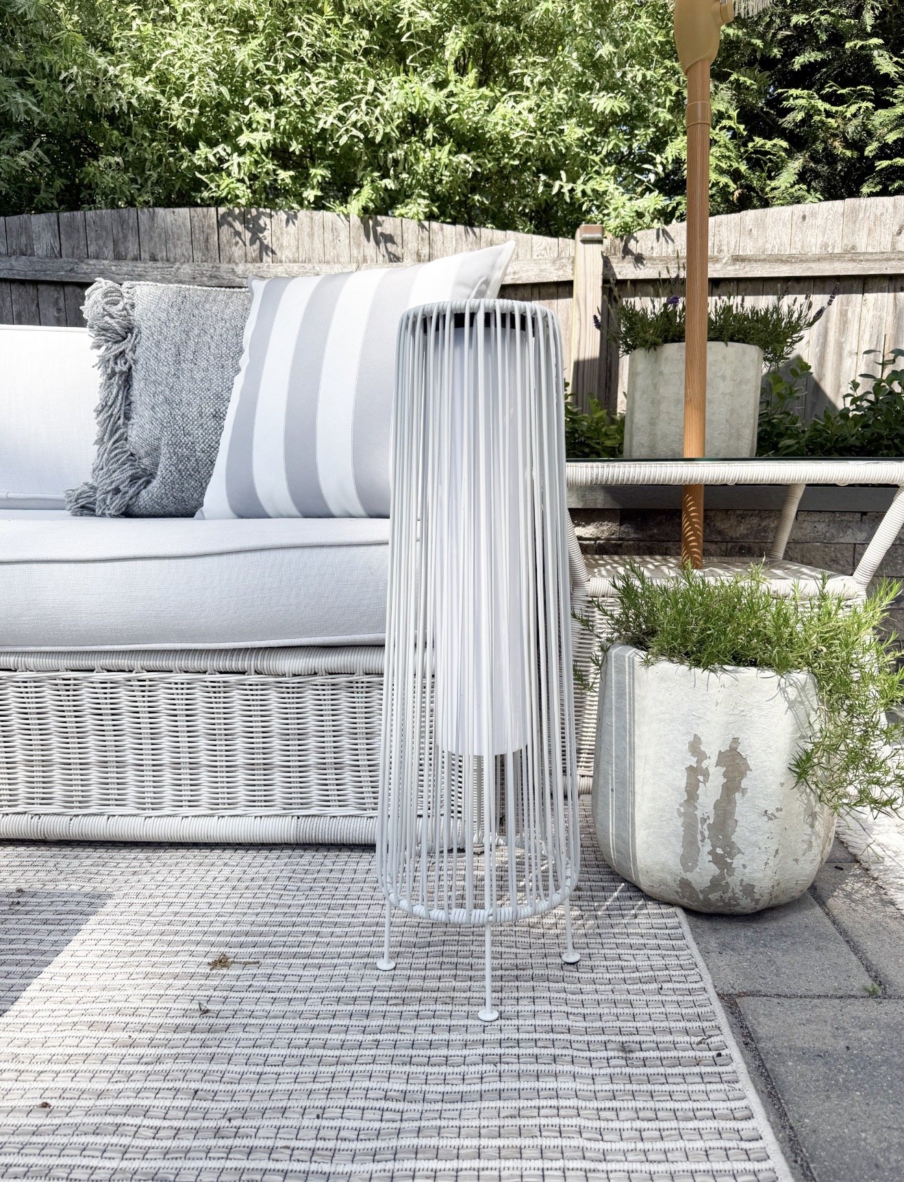 Modern solar floor lamp beside woven outdoor patio furniture, textured neutral pillows, and layered concrete planters on a cozy backyard patio with lush greenery.