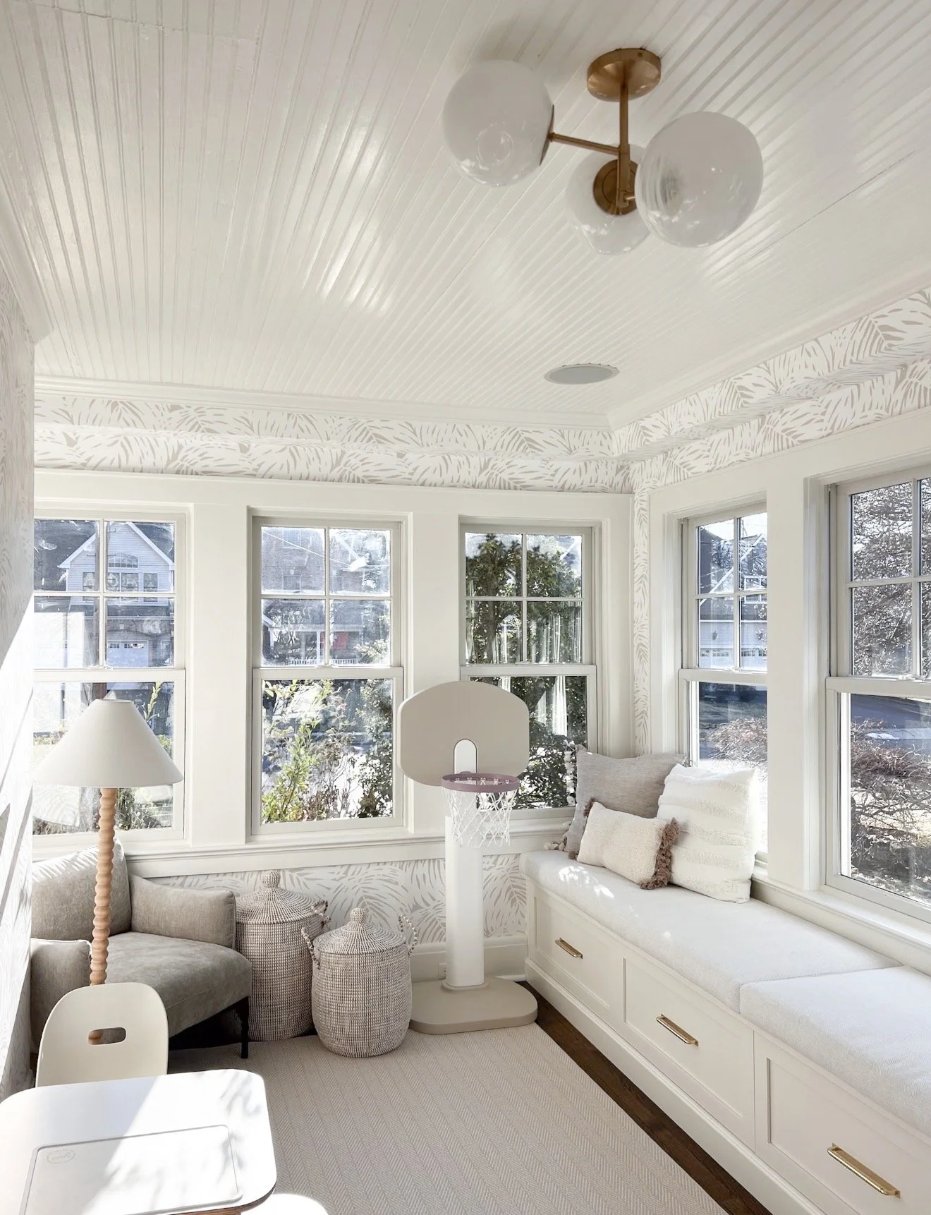 Bright sunroom with built in window seating, palm leaf wallpaper, beadboard ceiling, and brass globe ceiling light fixture.