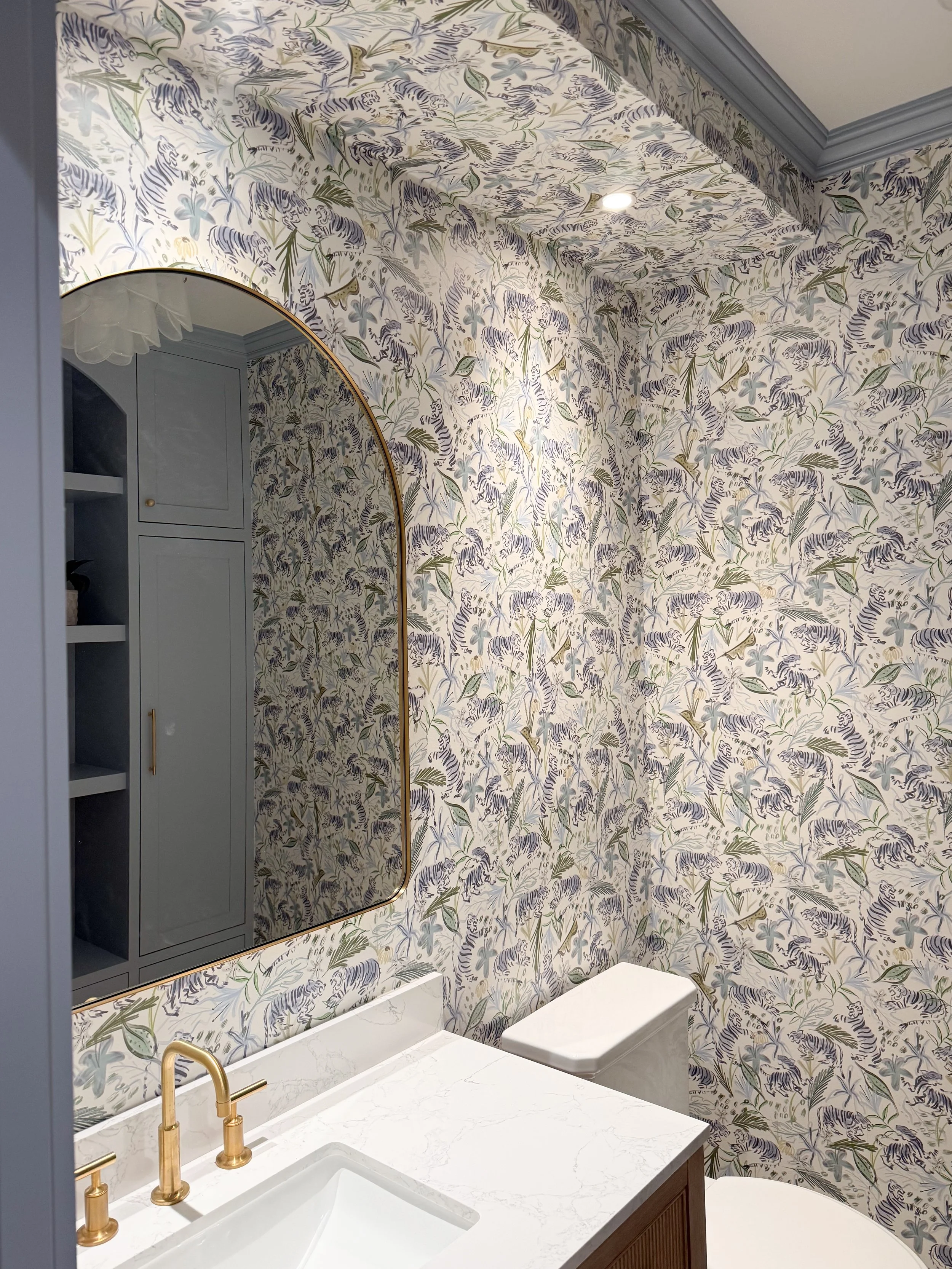 Client powder room with blue tiger wallpaper, wood vanity, marble countertop, brass mirror and brass fixtures.