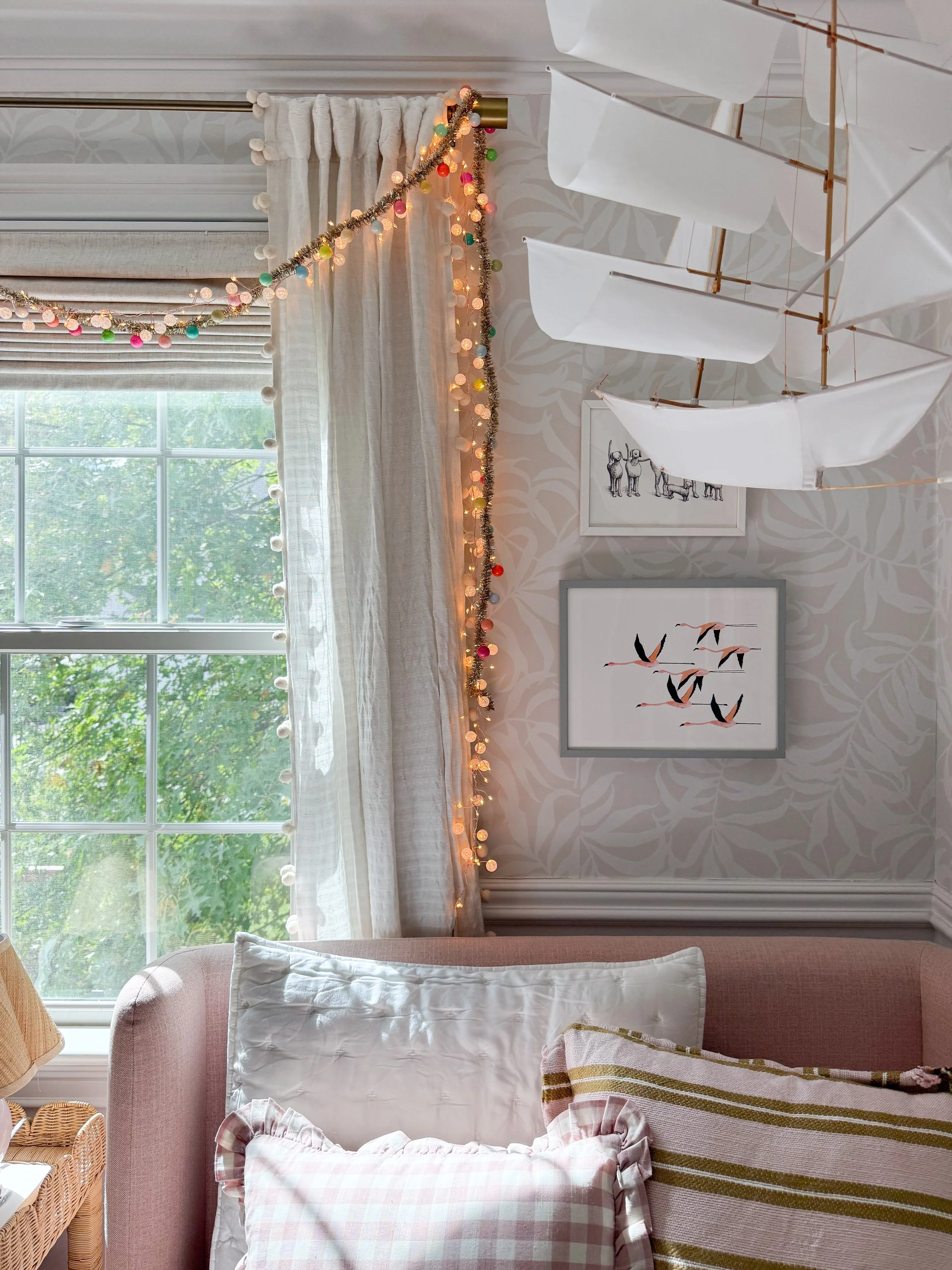 Anthropologie holiday bedroom decor featuring colorful Christmas string lights draped around cream curtains, a decorative white sailboat, framed artwork, and blush pink bedding with patterned pillows.