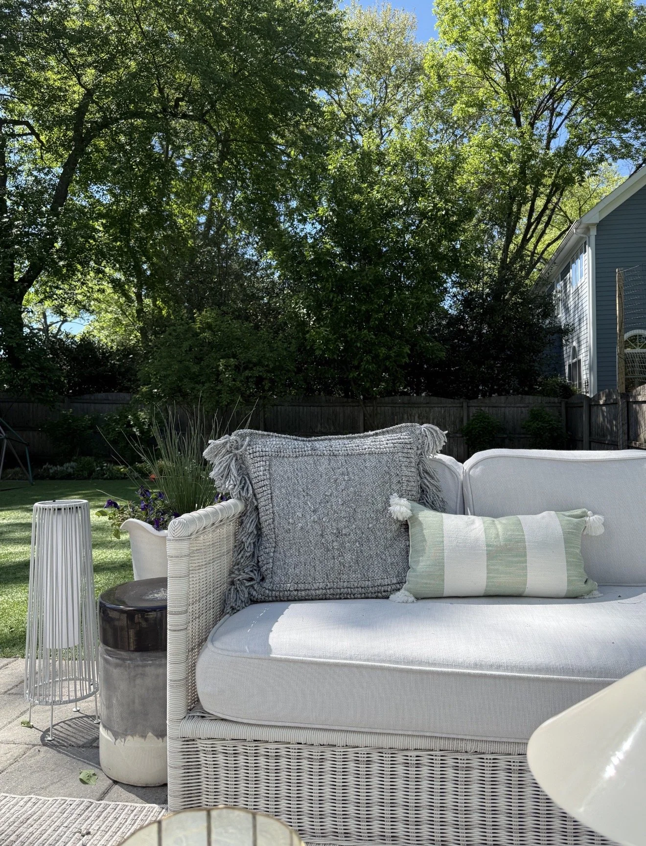Cozy outdoor lounge seating with woven patio furniture, textured neutral pillows, layered backyard landscaping, and mature trees creating a lush and relaxing outdoor retreat.