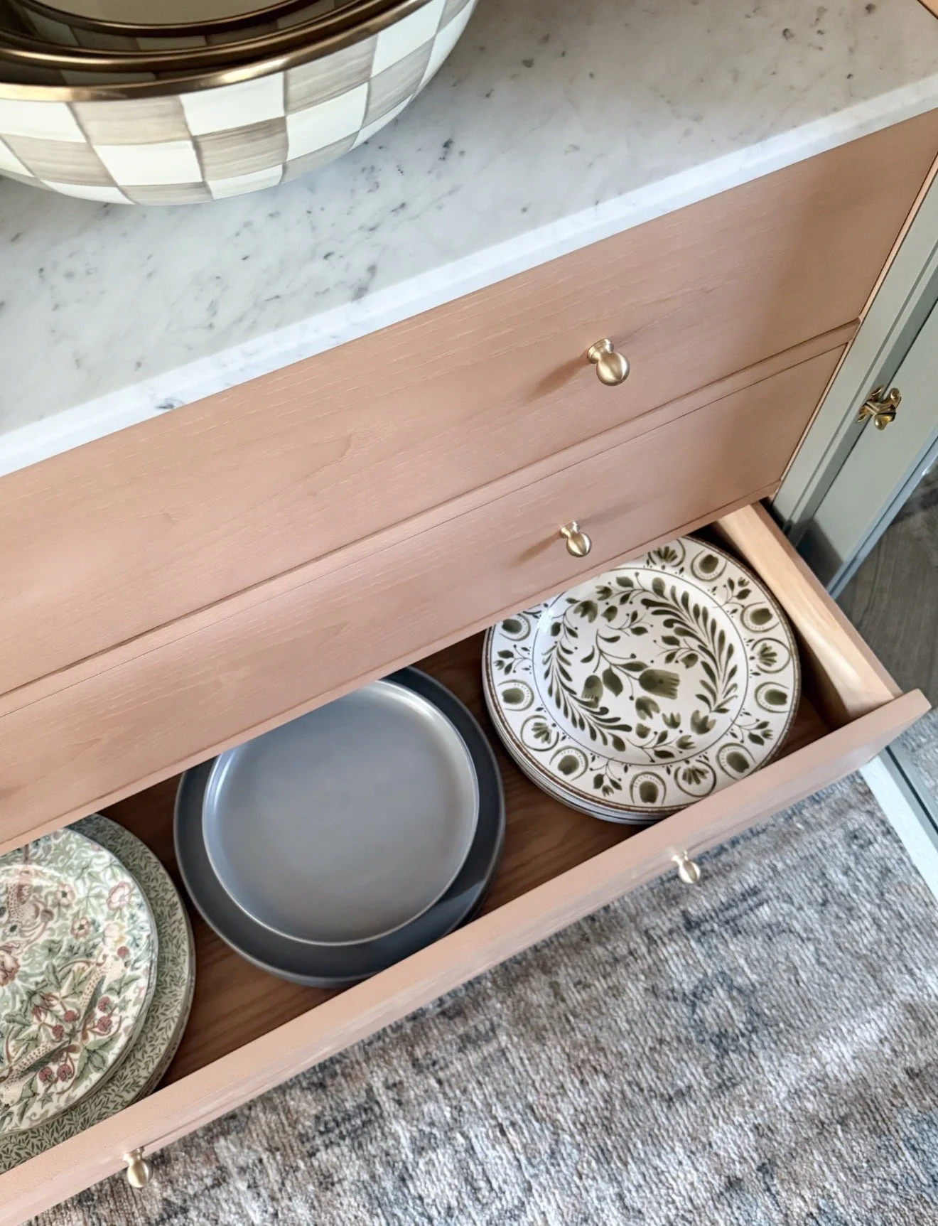 Close-up of a coral-toned built-in cabinet with brass knobs, open drawers displaying patterned plates, a marble countertop, and a neutral Loloi rug beneath.