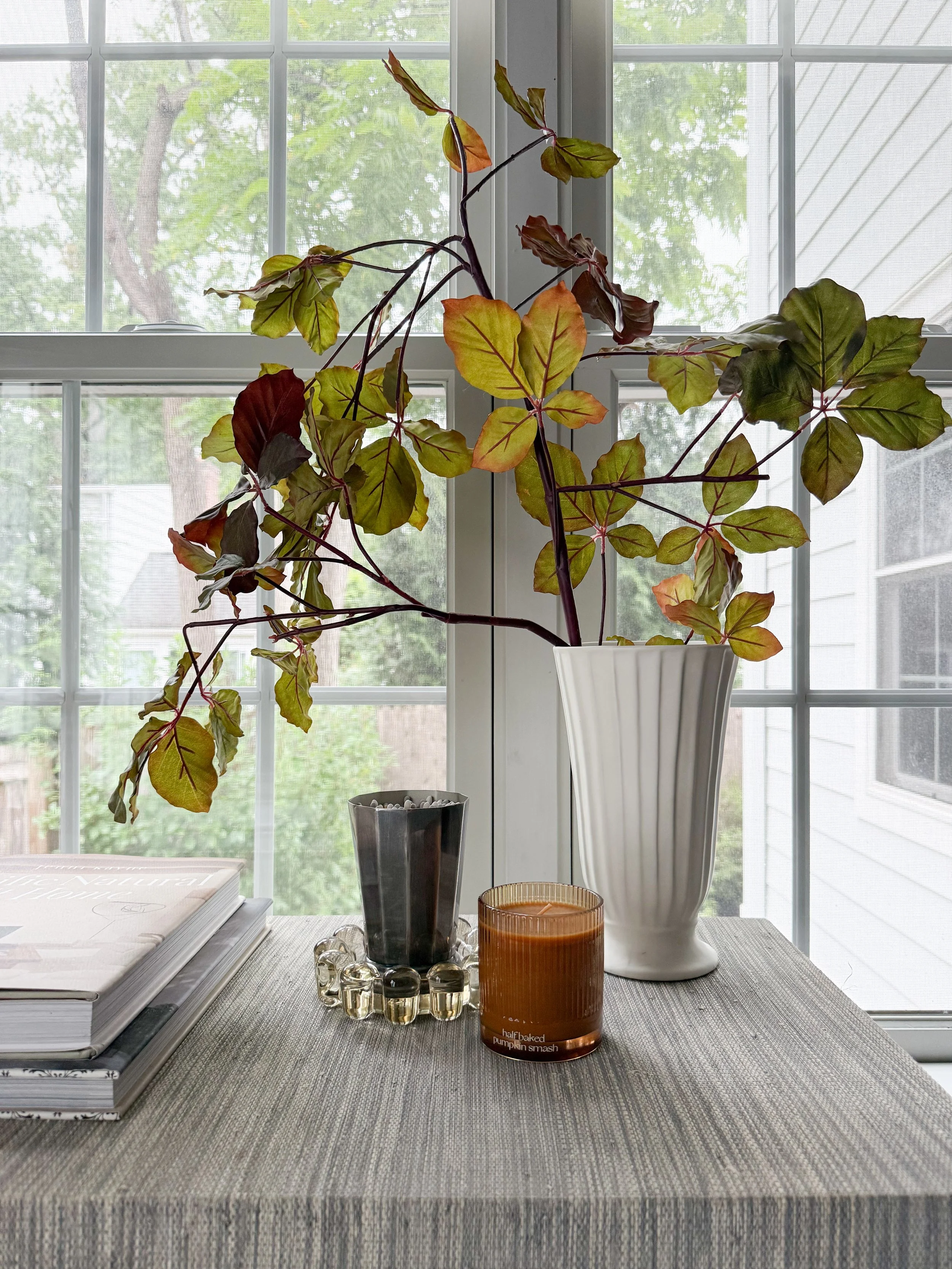 Autumn branches with olive green and rust leaves styled in a white vase beside an amber pumpkin candle for simple organic fall decor.