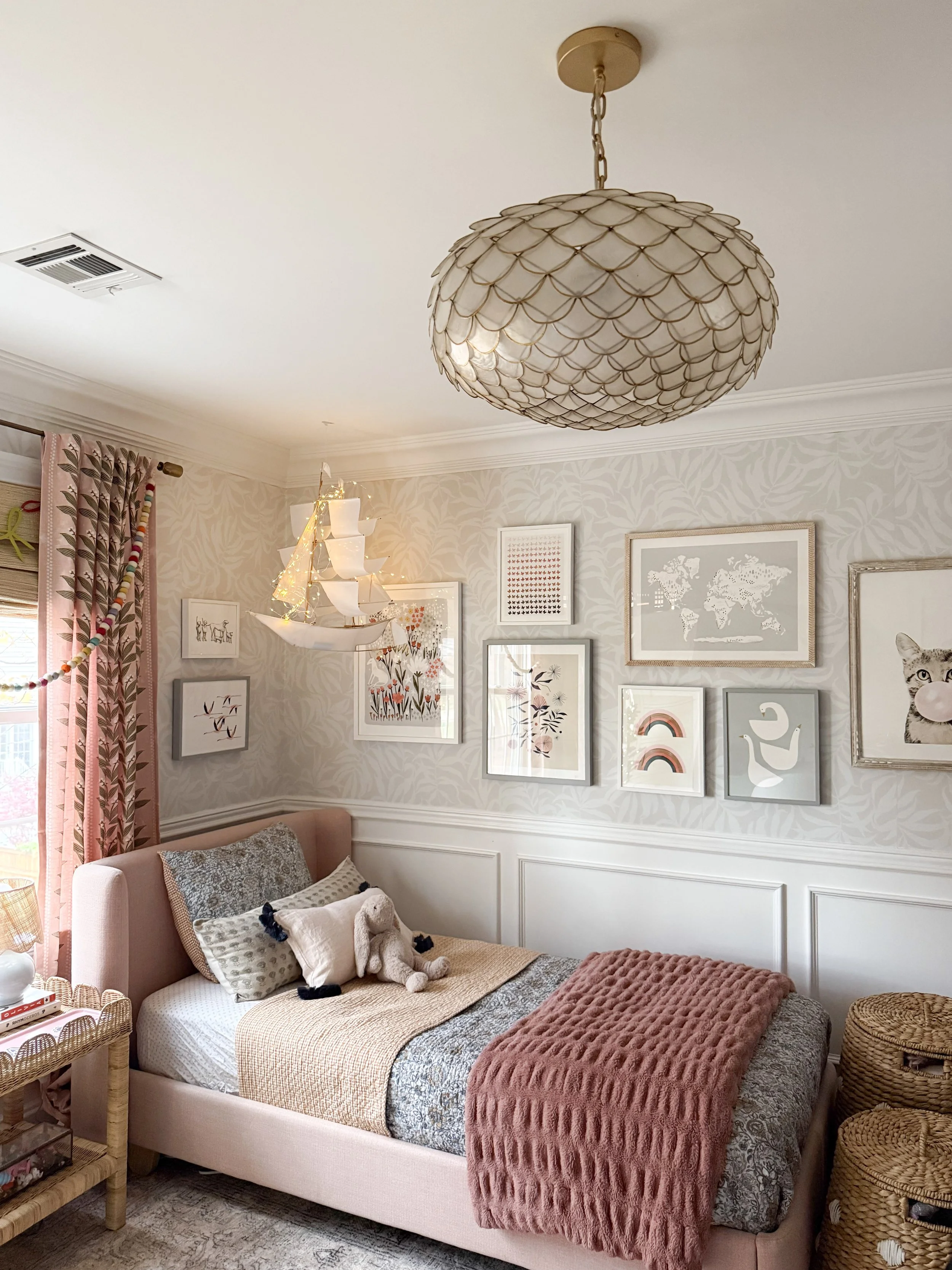 Girls bedroom with neutral botanical wallpaper, pink upholstered bed, gallery wall art, woven storage baskets, and warm layered bedding.
