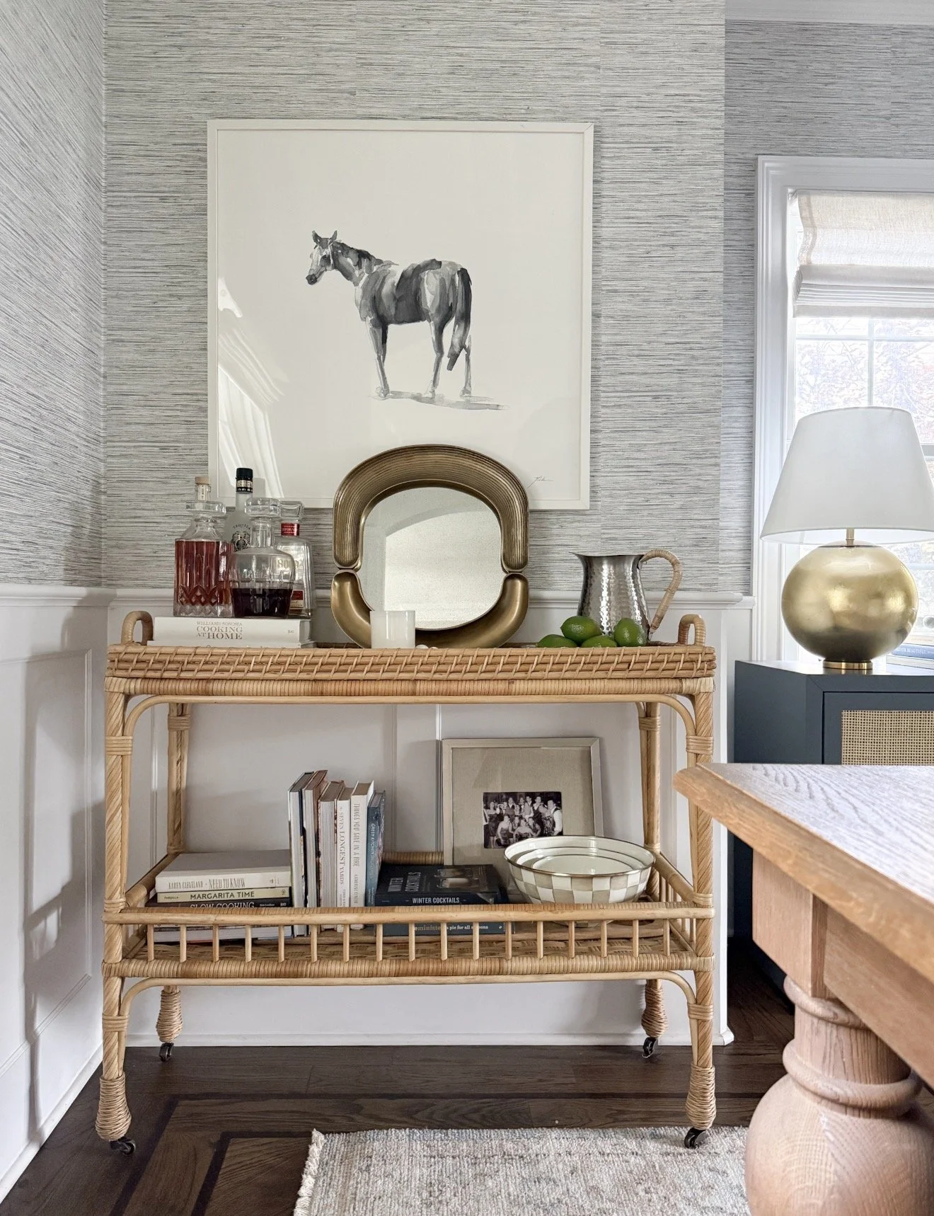 Woven rattan bar cart on casters styled with a round gold mirror, a pitcher, a gold lamp, liquor bottles, a small stack of books, and a decorative bowl.