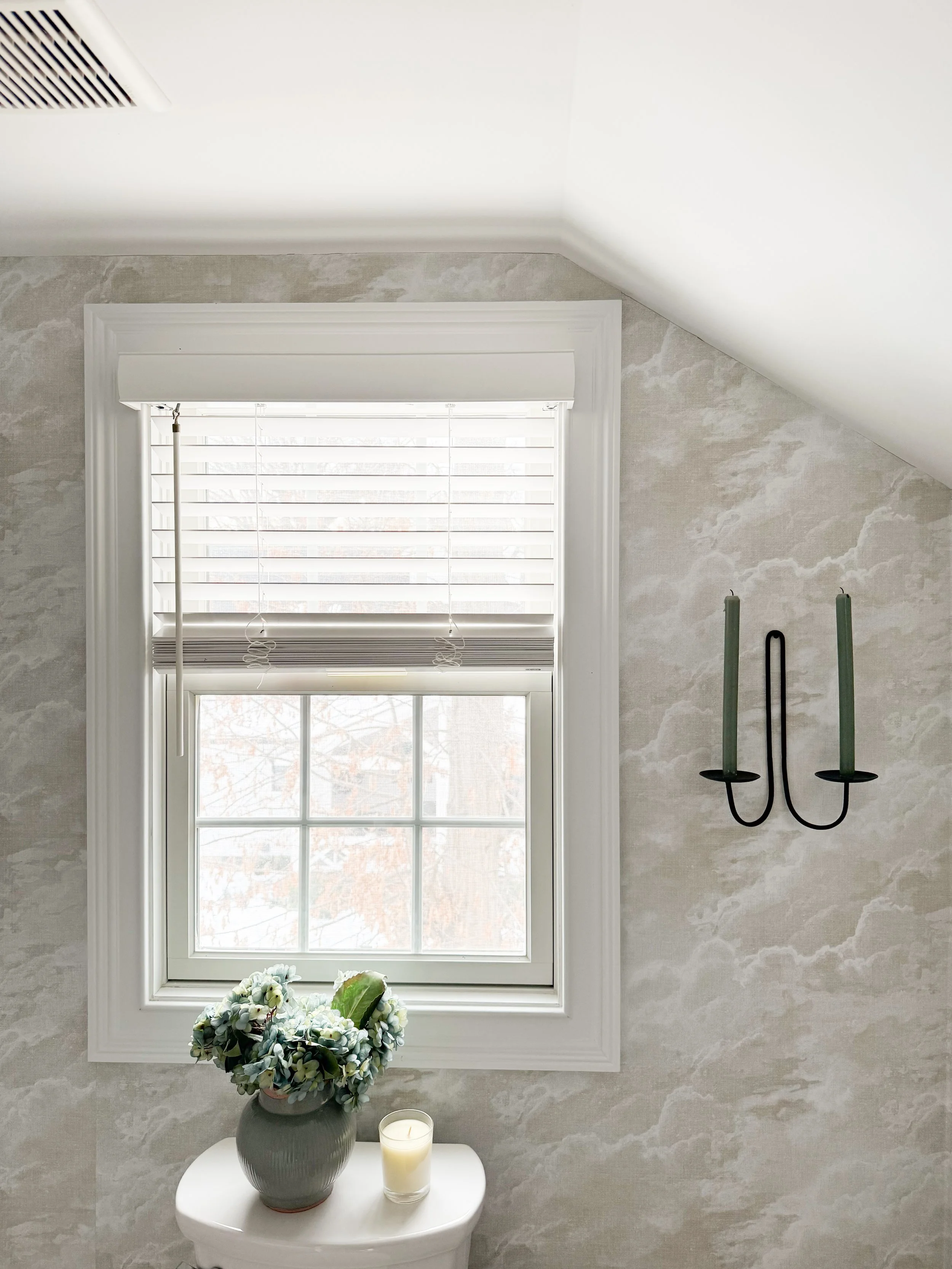 Bright bathroom window with white blinds above a toilet, styled with a vase of blue hydrangeas and a candle, set against textured wallpaper with a black wall sconce and sloped ceiling.