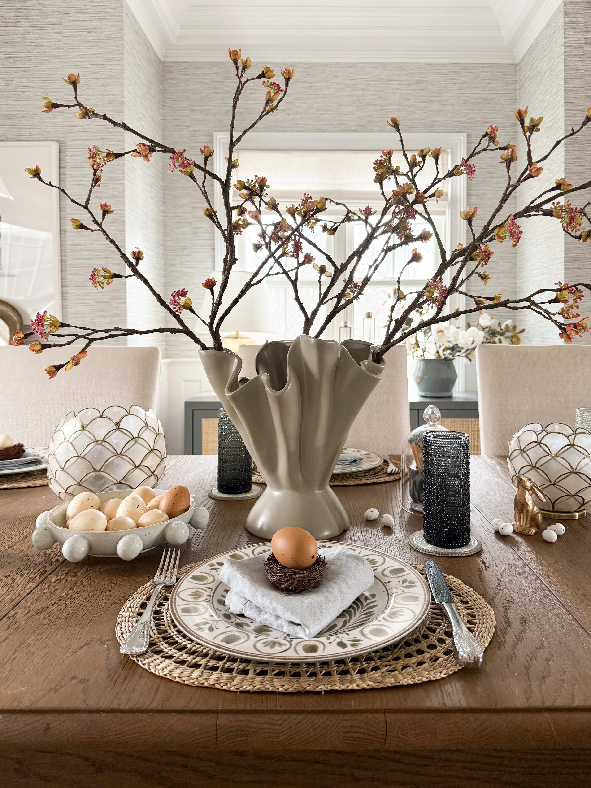 Neutral Easter tablescape detail with ceramic vase, decorative eggs, textured glass candle holders, and layered place setting on wood table