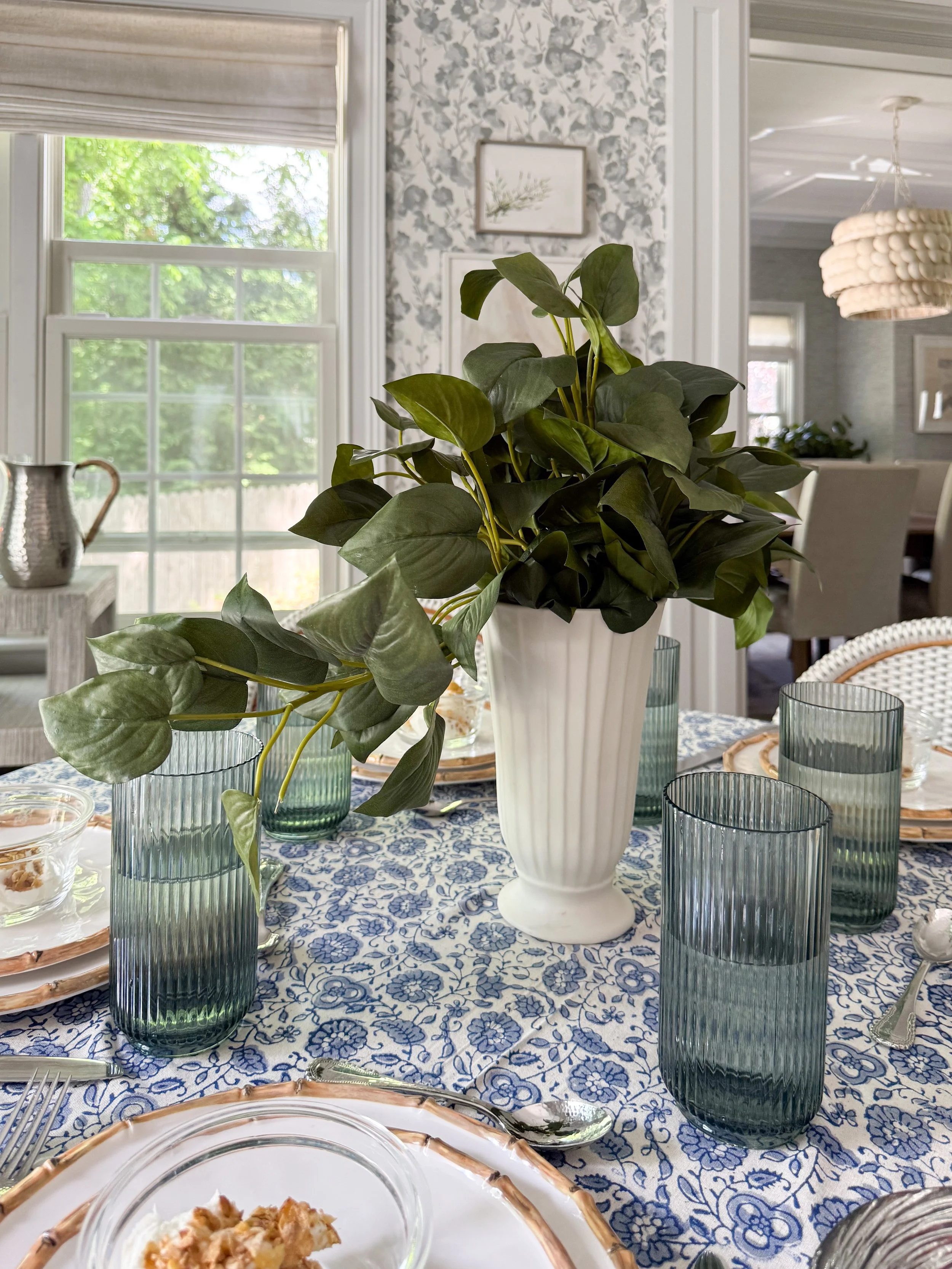 Overhead view of a McGee & Co inspired dining table with blue and white tablecloth, layered place settings, woven chargers, fresh greenery centerpiece, and summer entertaining decor.