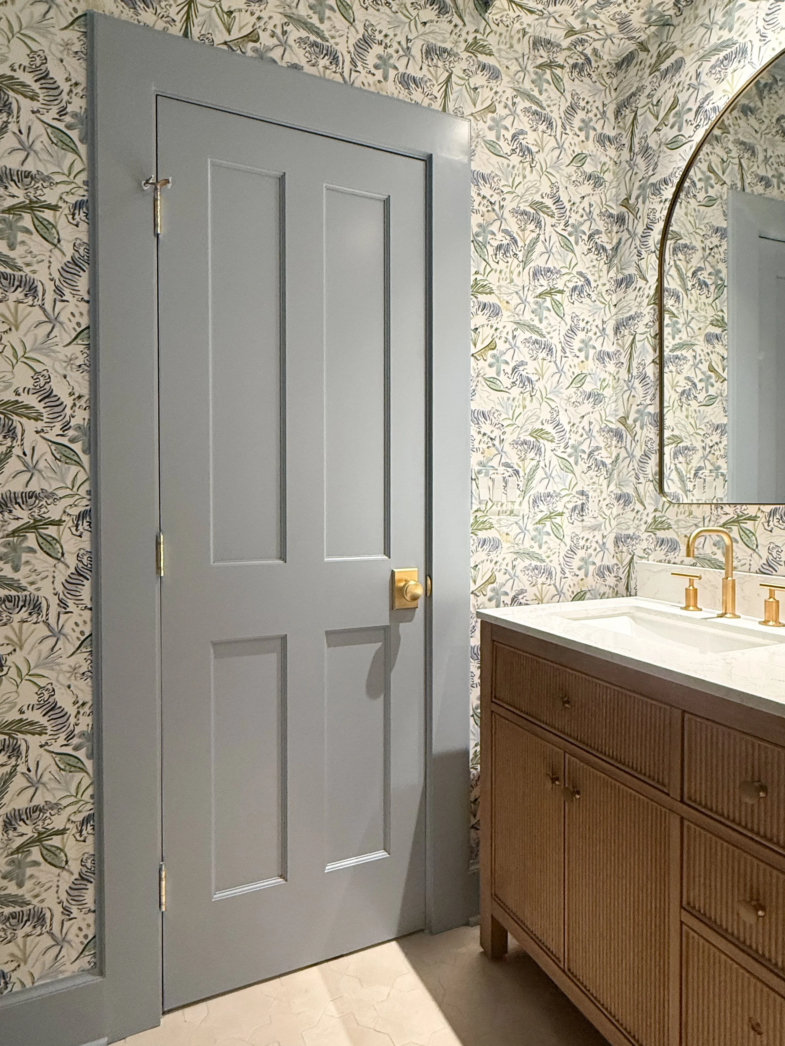 View into a colorful client powder room featuring blue tiger wallpaper, natural wood vanity and brass accents.