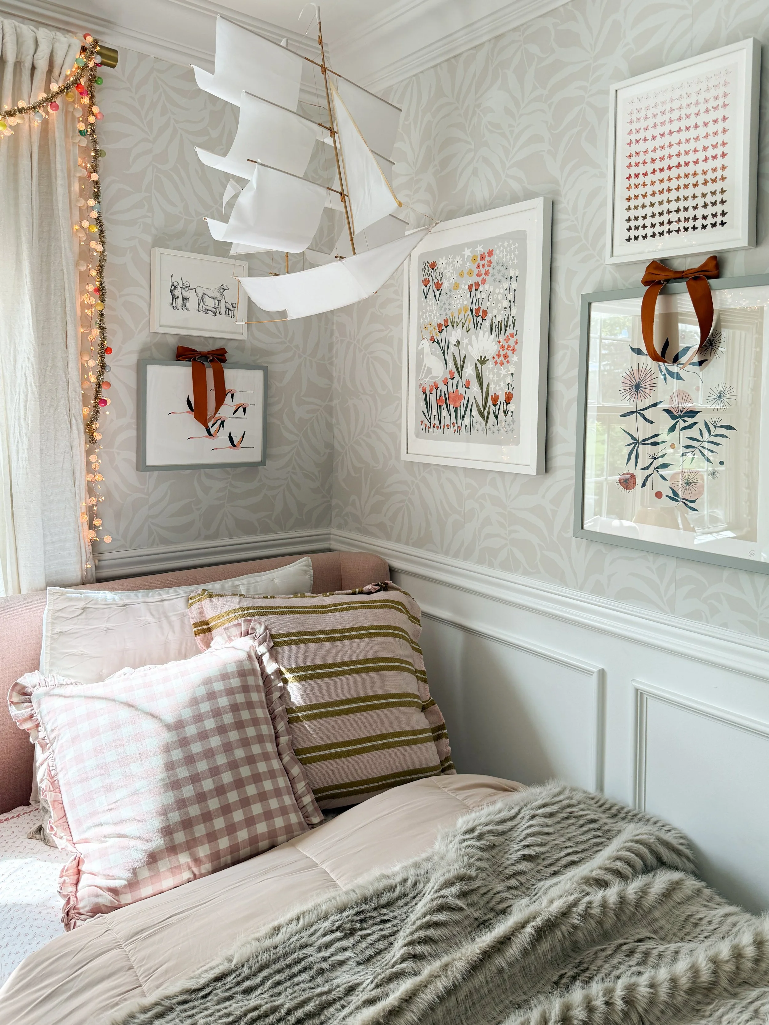 Anthropologie holiday bedroom decor featuring colorful framed artwork with ribbon accents, a white decorative sailboat, multicolored string lights, blush pink gingham pillows, and a cozy faux fur throw.