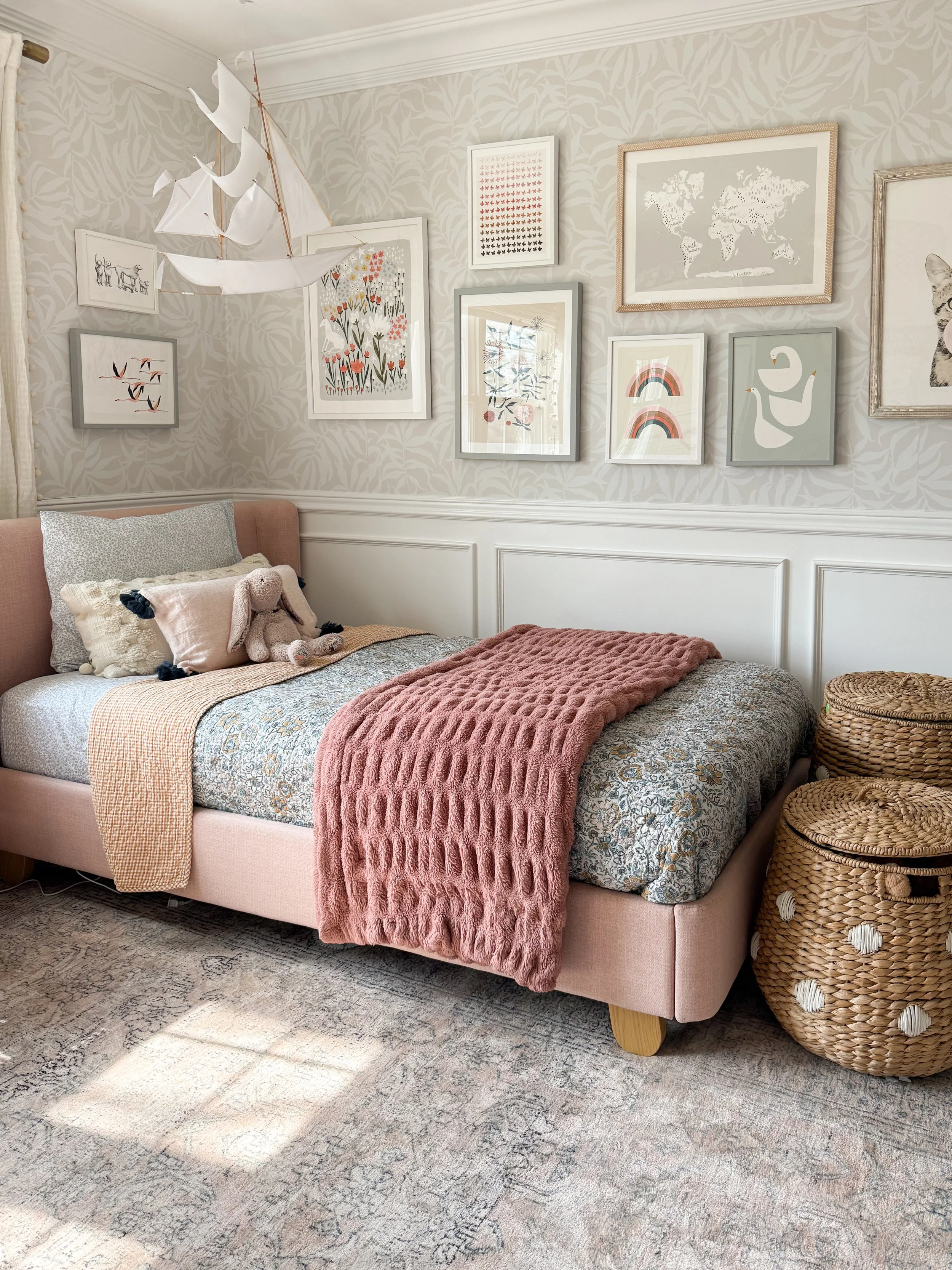 Girl's bedroom with a blush upholstered bed, neutral vintage style rug, gallery wall, and soft pink accents.