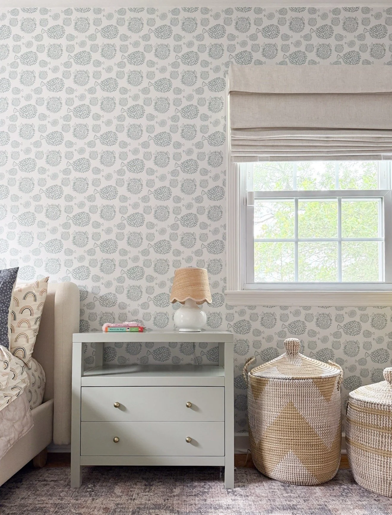 Room with soft blue patterned wallpaper, pink geometric curtain panels, neutral ottoman, and woven basket.