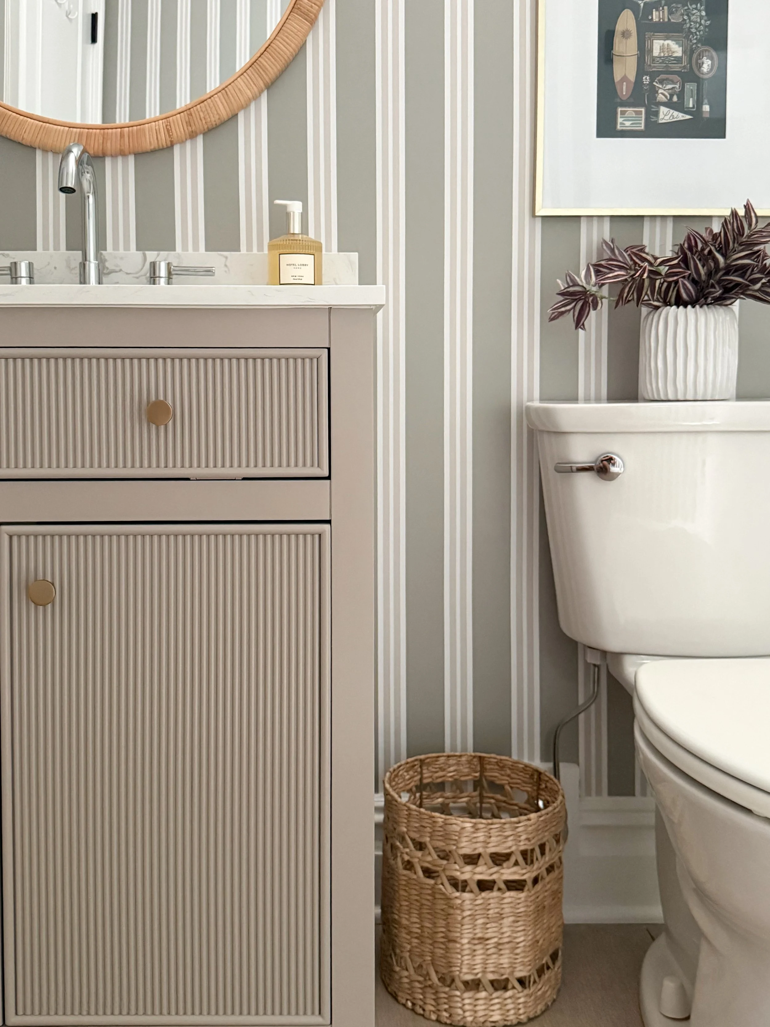 Basement bathroom design featuring Chasing Paper striped wallpaper, fluted vanity, rattan mirror, woven basket, framed artwork, and neutral decor accents.