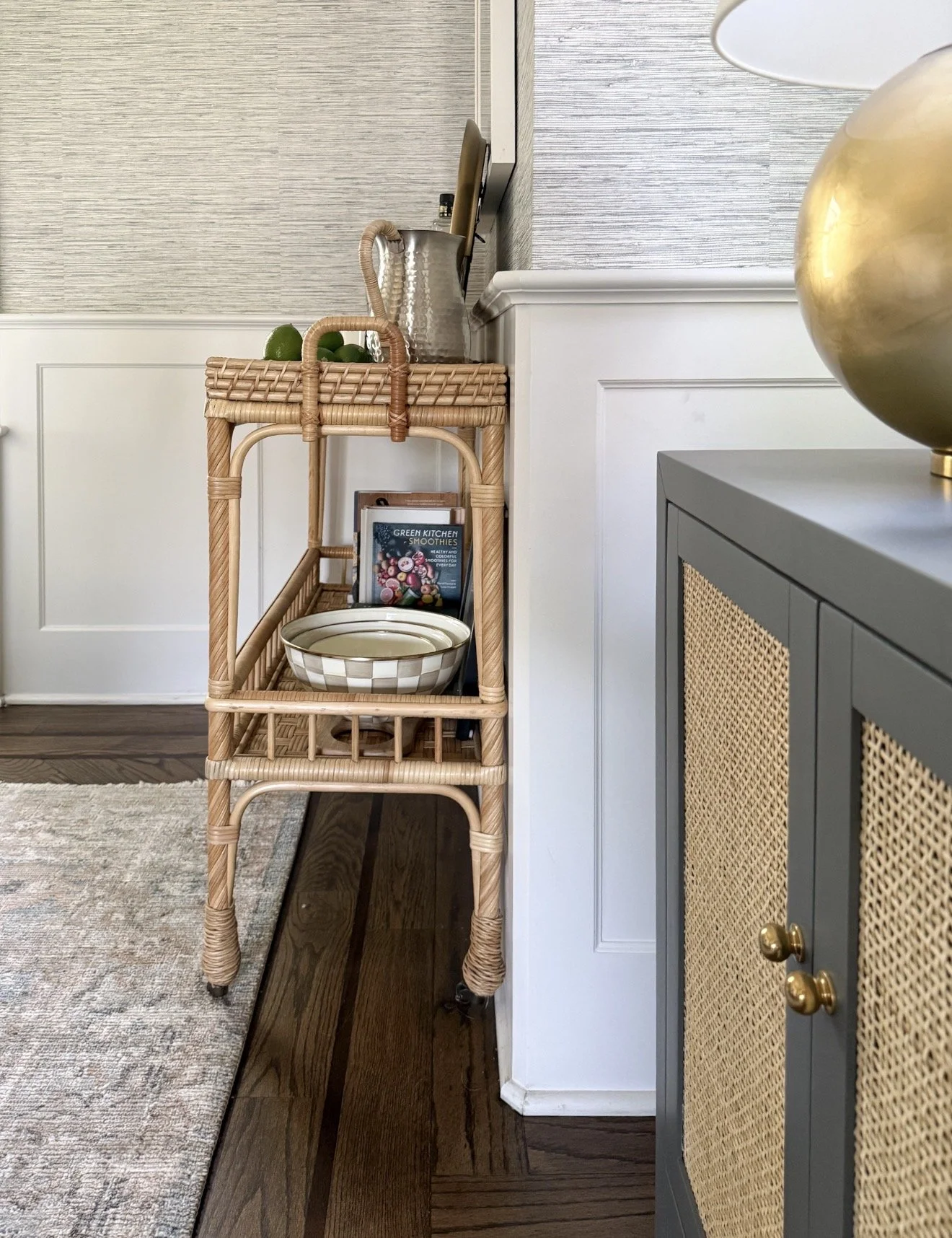 Woven rattan bar cart on casters styled with a round gold mirror, a pitcher, a gold lamp, liquor bottles, a small stack of books, and a decorative bowl.