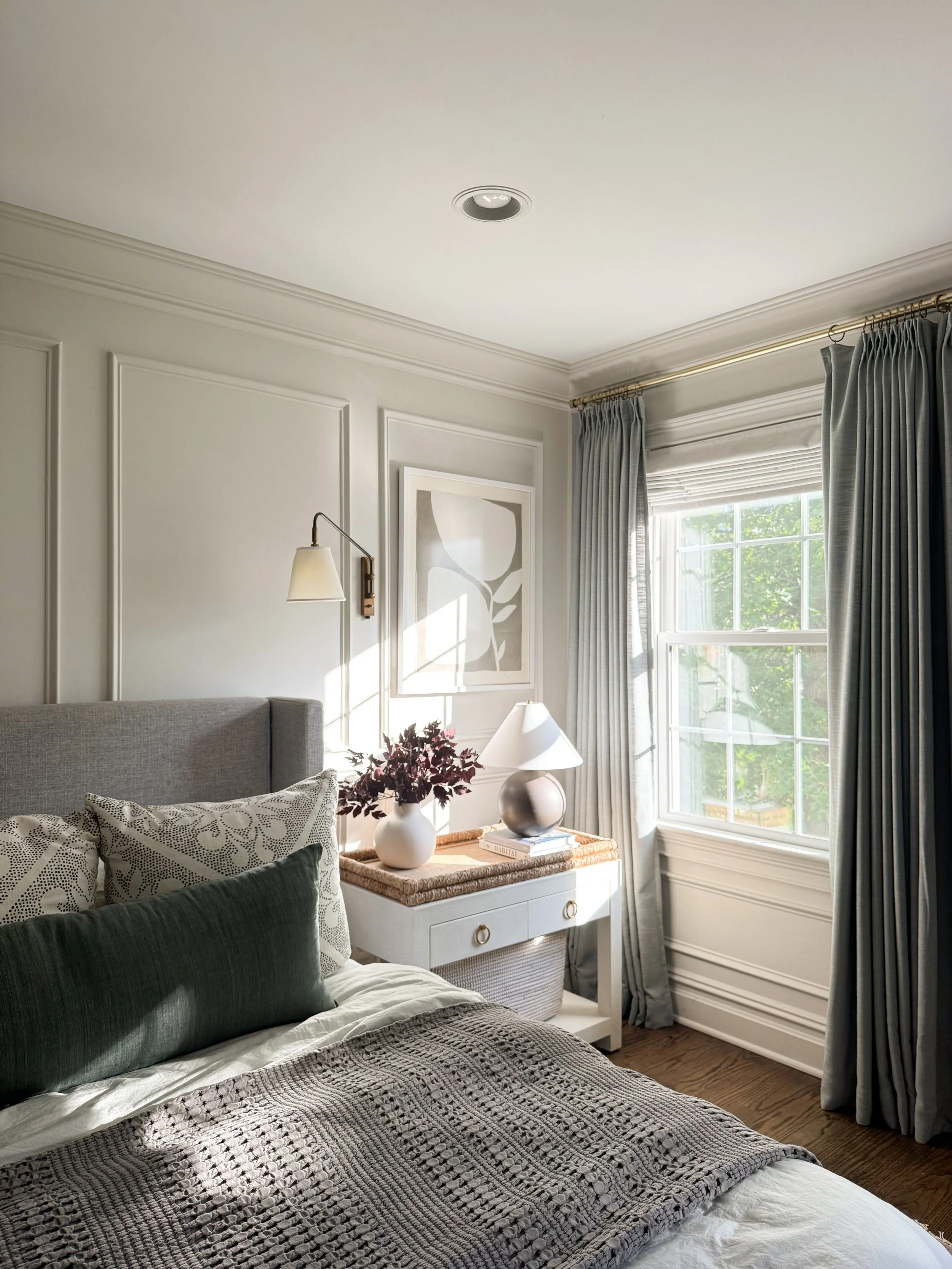 Cozy neutral bedroom styled for fall with layered bedding, a textured knit throw, deep green accent pillow, autumn branches, and soft gray curtains.