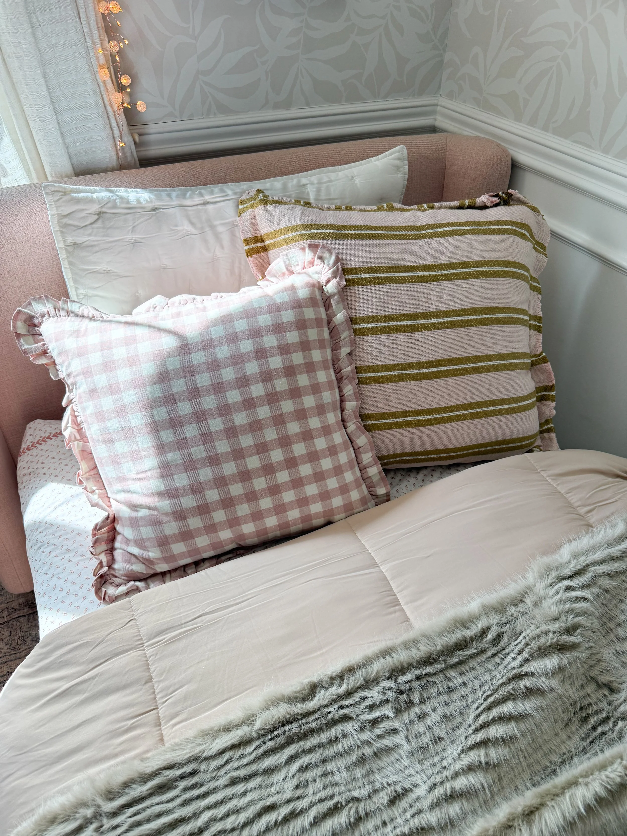 Anthropologie holiday bedroom decor featuring blush pink gingham pillows, an olive green striped pillow, neutral bedding, and a plush faux fur throw layered on a cozy bed.