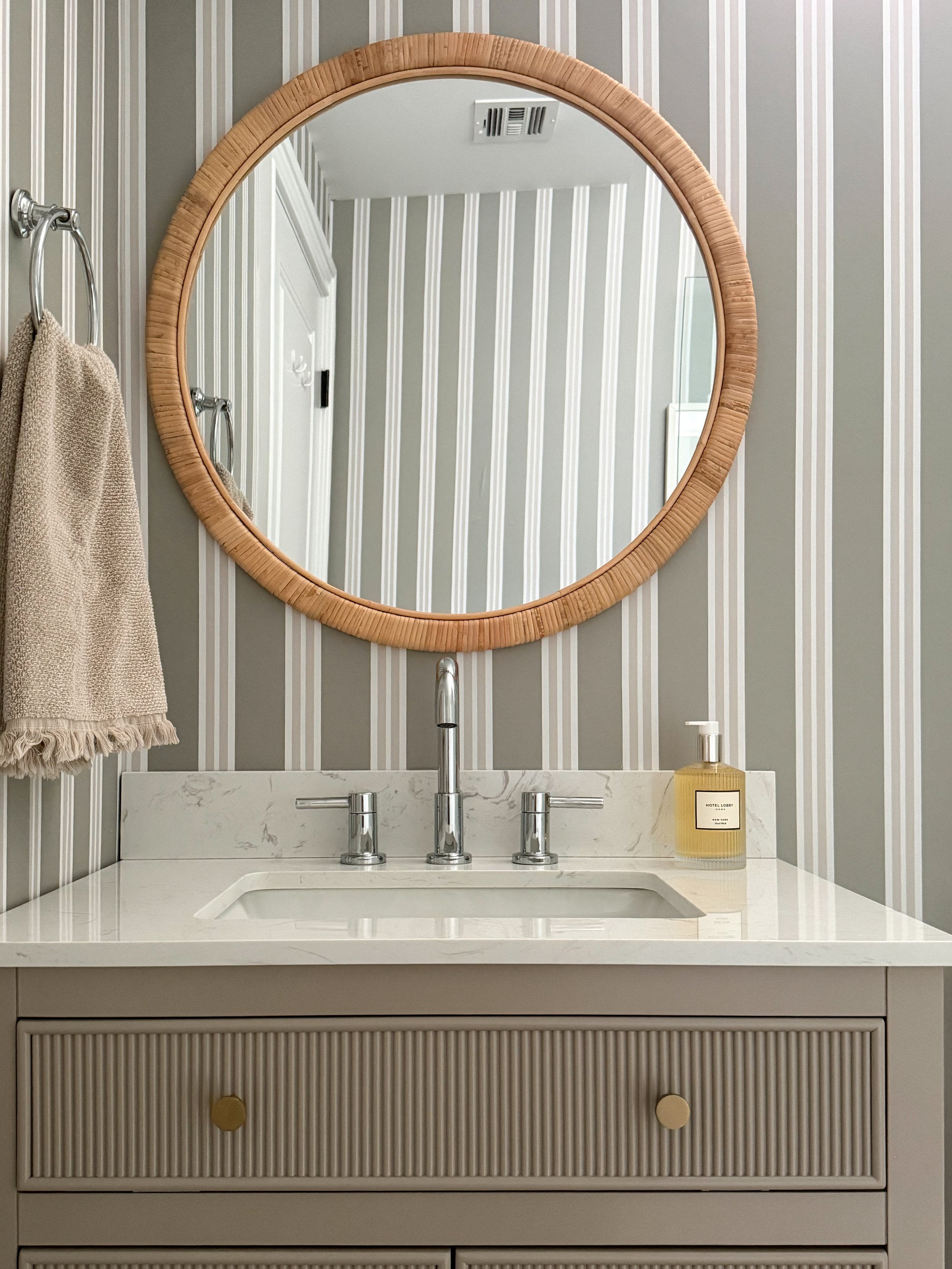 Basement bathroom vanity with Chasing Paper striped wallpaper, rattan round mirror, marble countertop, chrome fixtures, and neutral styling details for a small bathroom makeover.