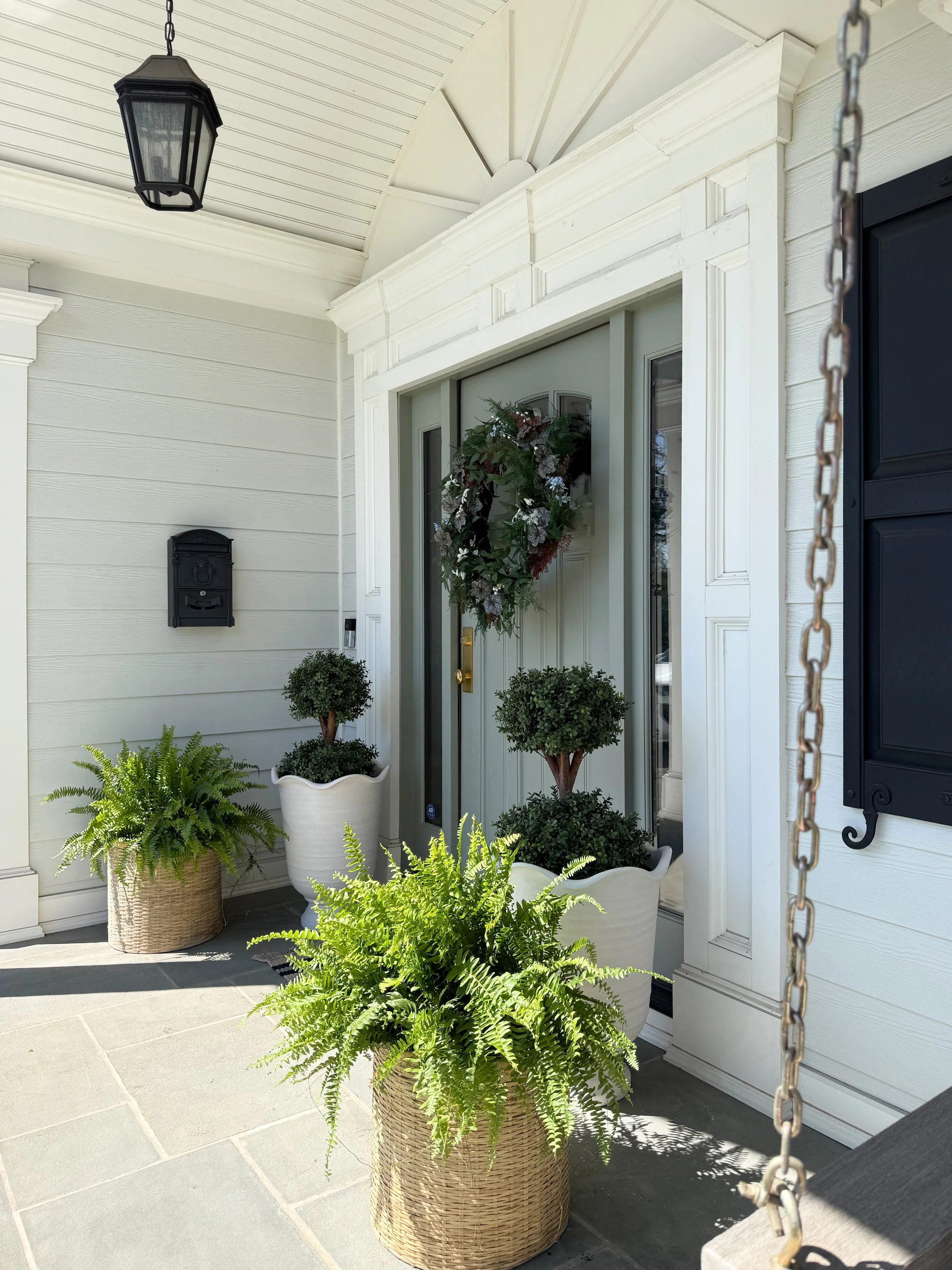 Angled view of front porch with layered planters, greenery, black lantern light, and classic entry details