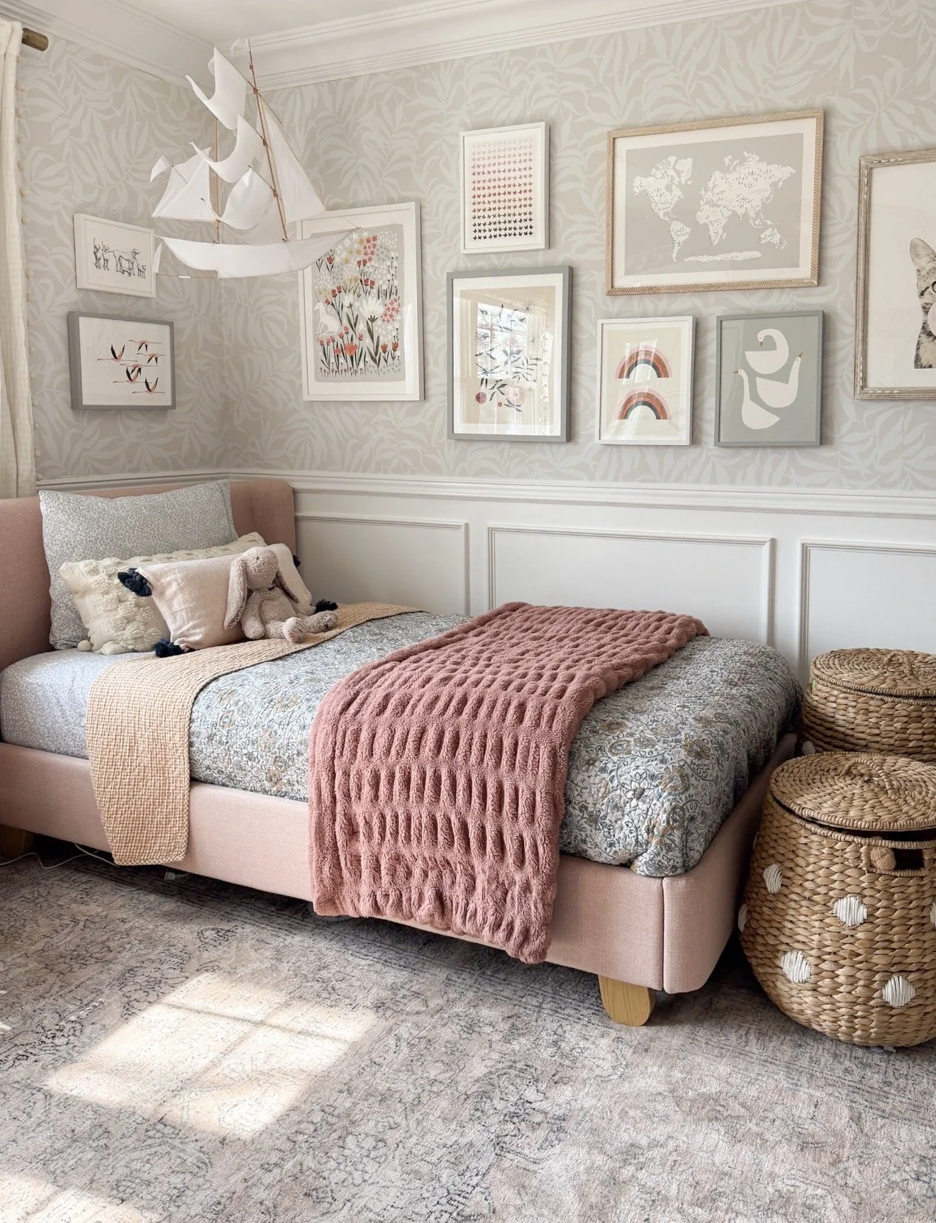 Close-up view of a muted blue and beige patterned area rug under the corner of a bed with white and gray bedding, showing the detailed floral and geometric design of the rug in a bedroom setting.