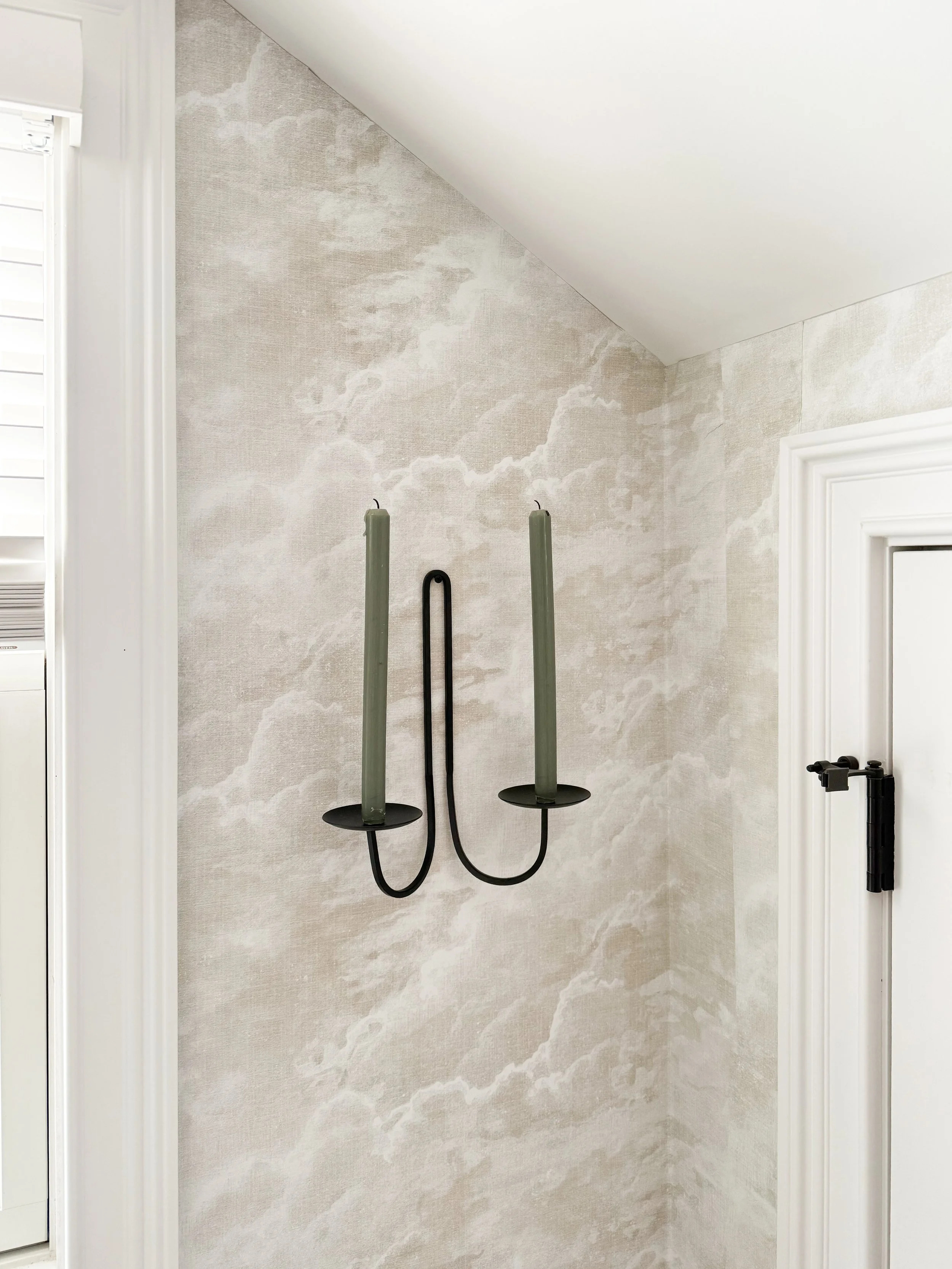 Black wall mounted double candle sconce with green taper candles, styled against soft textured neutral wallpaper in a corner with white trim and a sloped ceiling.