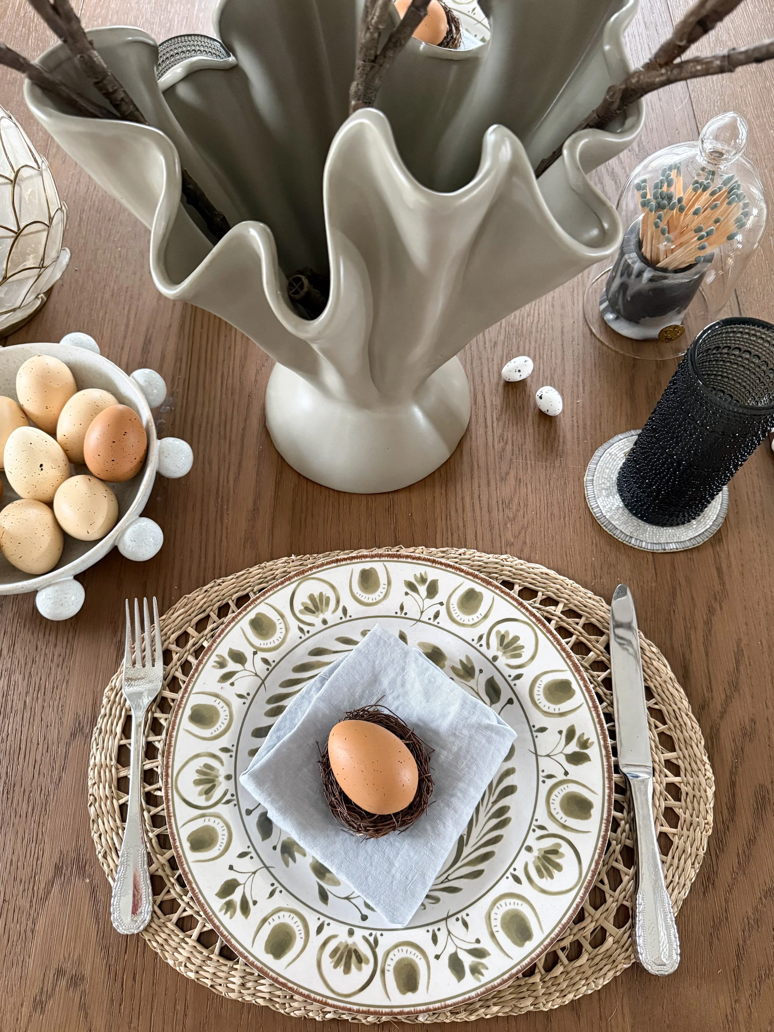Close up of spring table decor with ceramic vase, speckled Easter eggs in decorative bowl, and textured candle holders on wood dining table