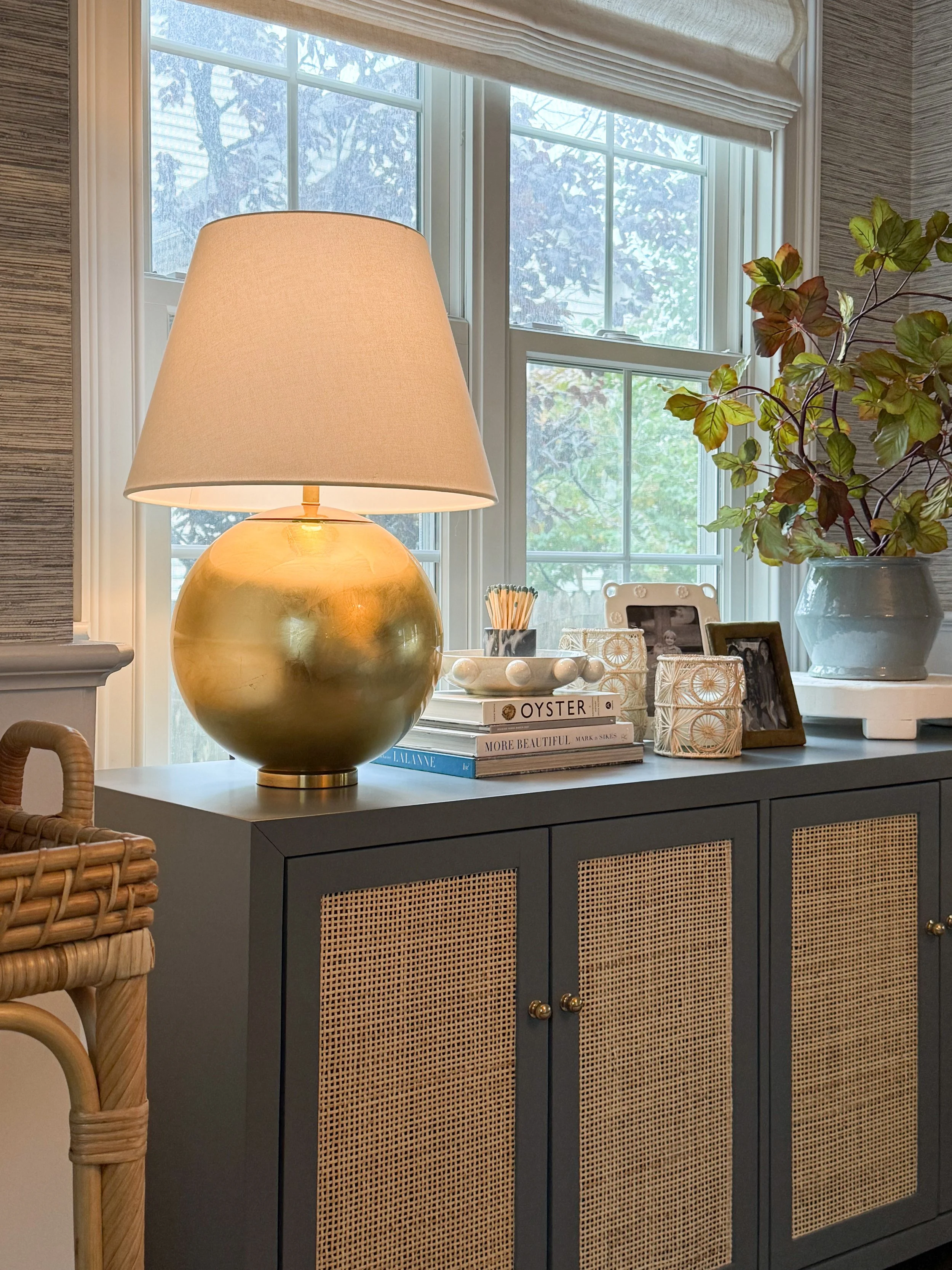 Brass table lamp with a white linen shade styled on a dining room cabinet with decorative accessories.