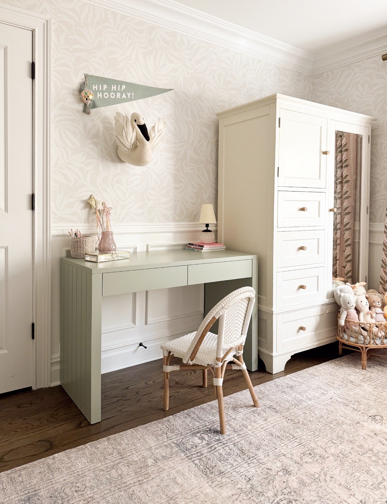 A light green Crate & Kids Parsons desk in a child’s bedroom next to a white storage cabinet. The desk is styled with a small lamp, pink vase, pencil holder, and tray, and paired with a white and natural woven chair.