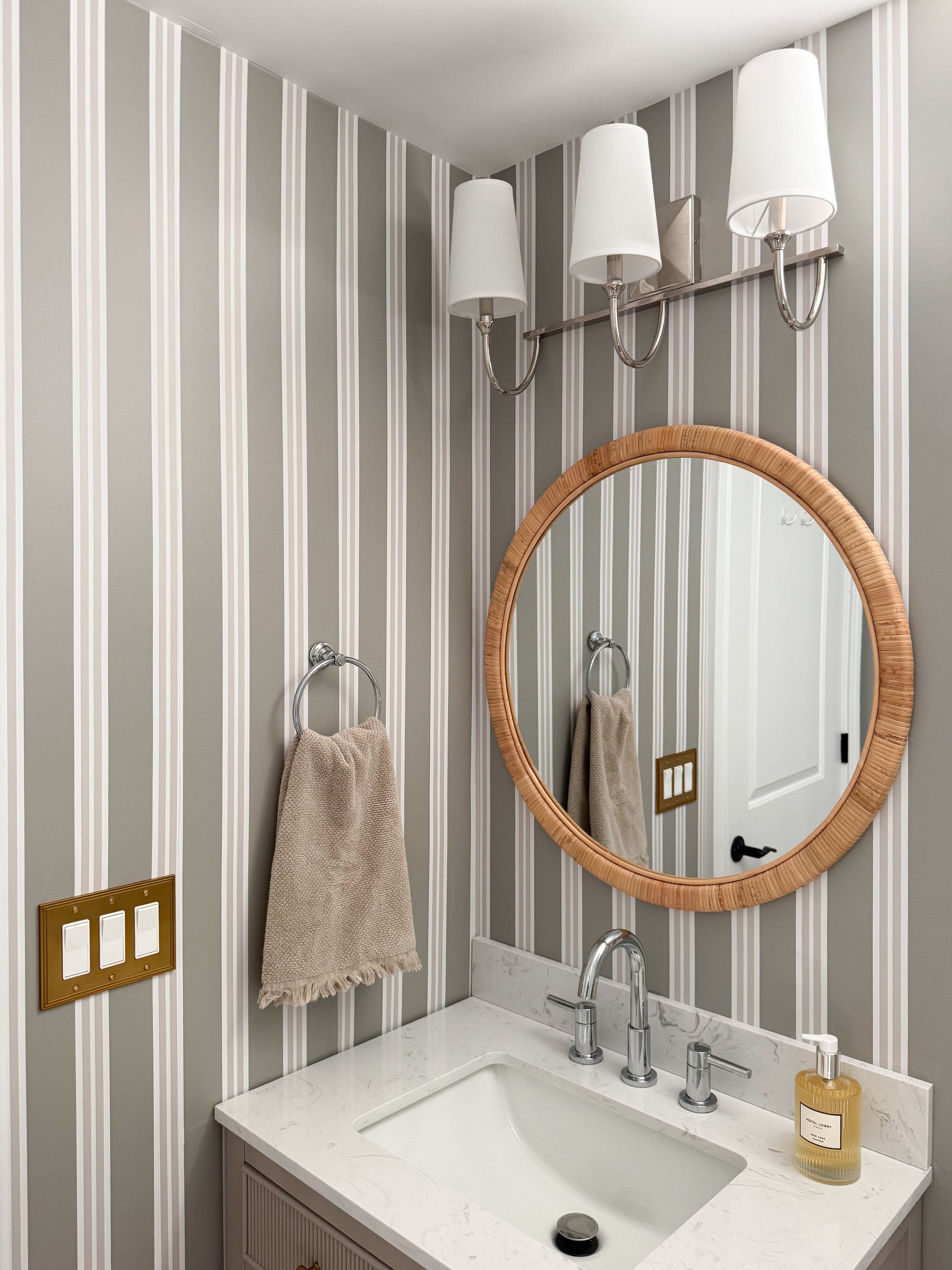 Basement bathroom makeover featuring Chasing Paper striped wallpaper, a rattan round mirror, marble vanity countertop, chrome fixtures, and neutral bathroom decor.