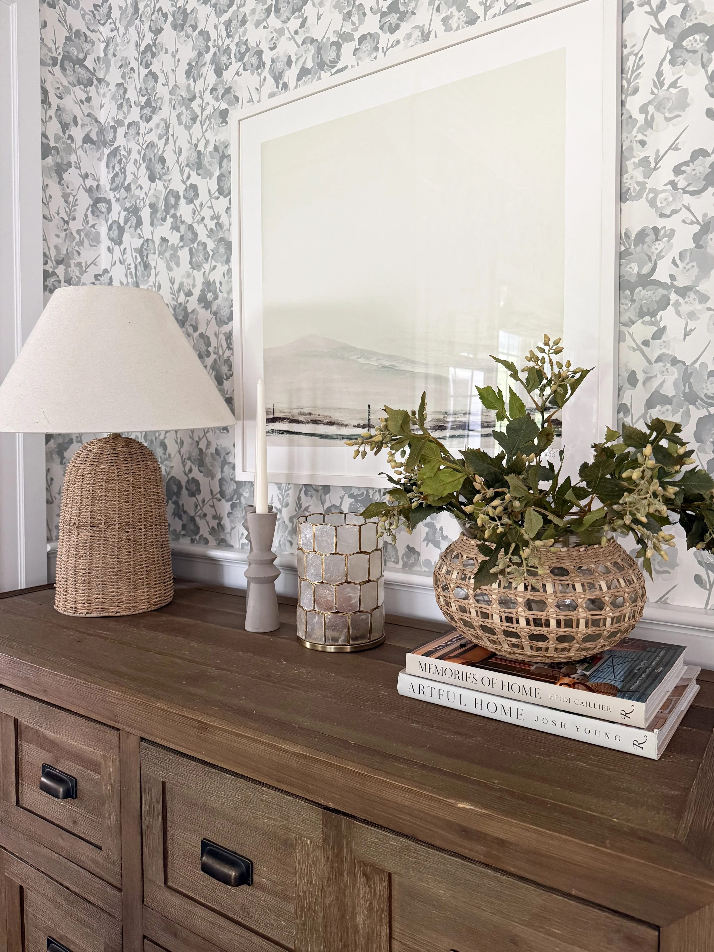 Warm wood console styled with a woven table lamp, greenery in a rattan vase, stacked design books, candles, and soft blue floral wallpaper.