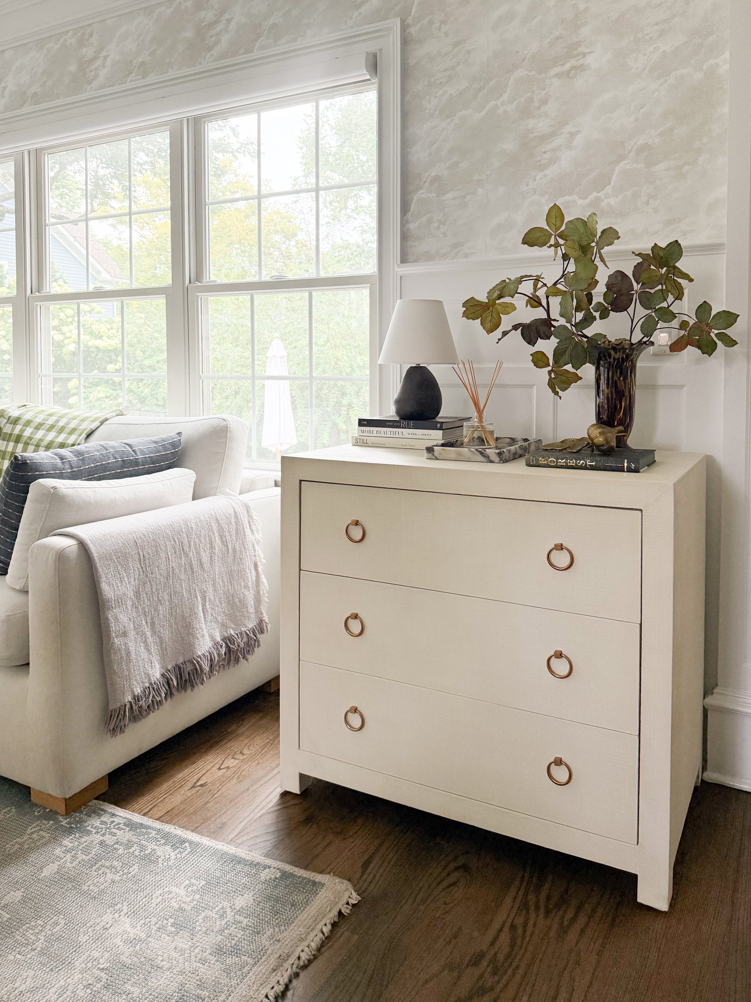 Neutral living room corner styled for fall with autumn branches in a dark vase, stacked books, a table lamp, and warm organic accents.