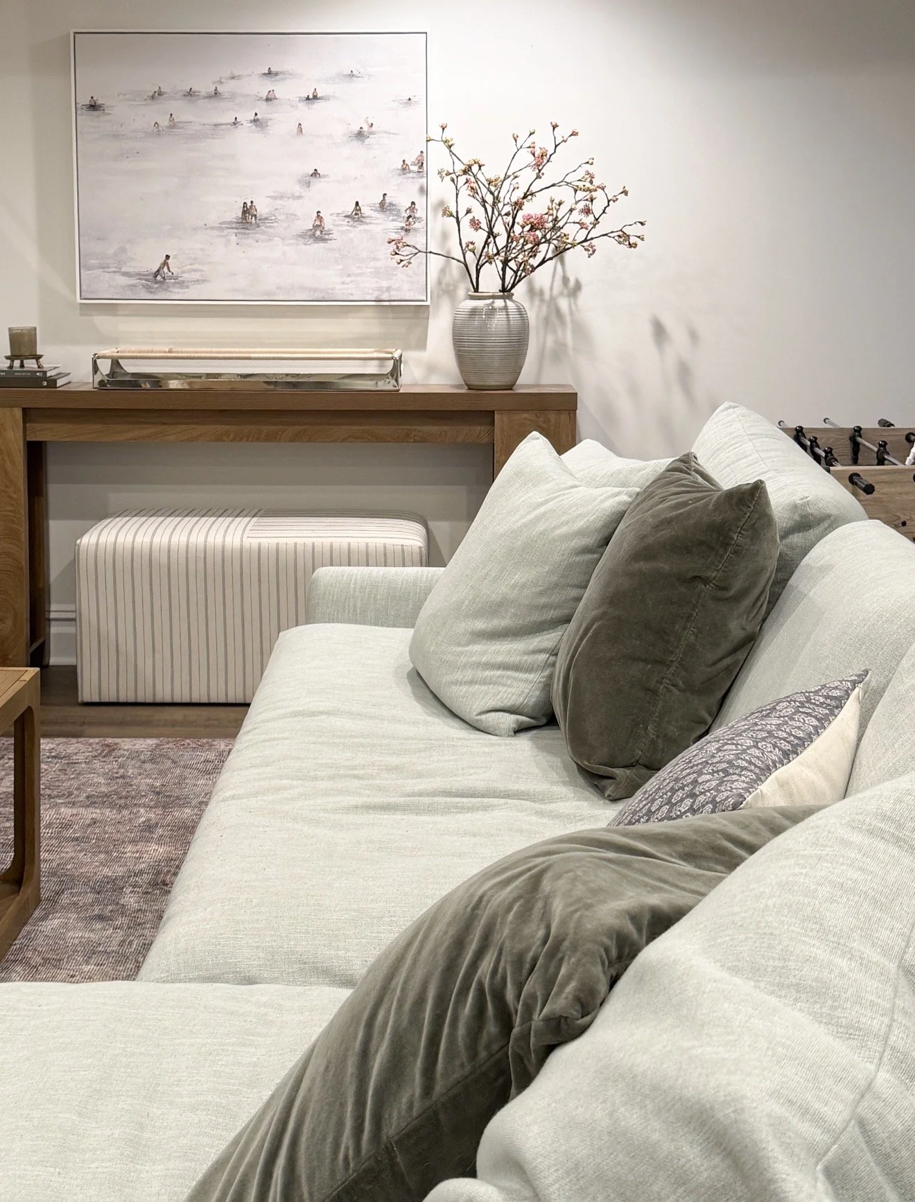 Modern basement living room with a soft sage green sectional sofa, velvet and patterned pillows, a light wooden coffee table, a gray accent chair, a striped ottoman, and a foosball table in the background.