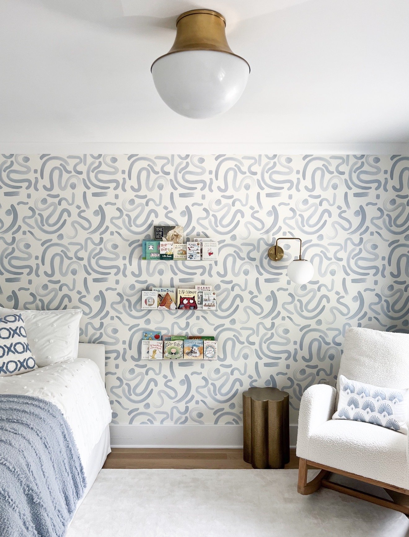 Children's bedroom with blue patterned wallpaper, brass wall sconce, neutral flush mount ceiling light, and cozy reading corner.