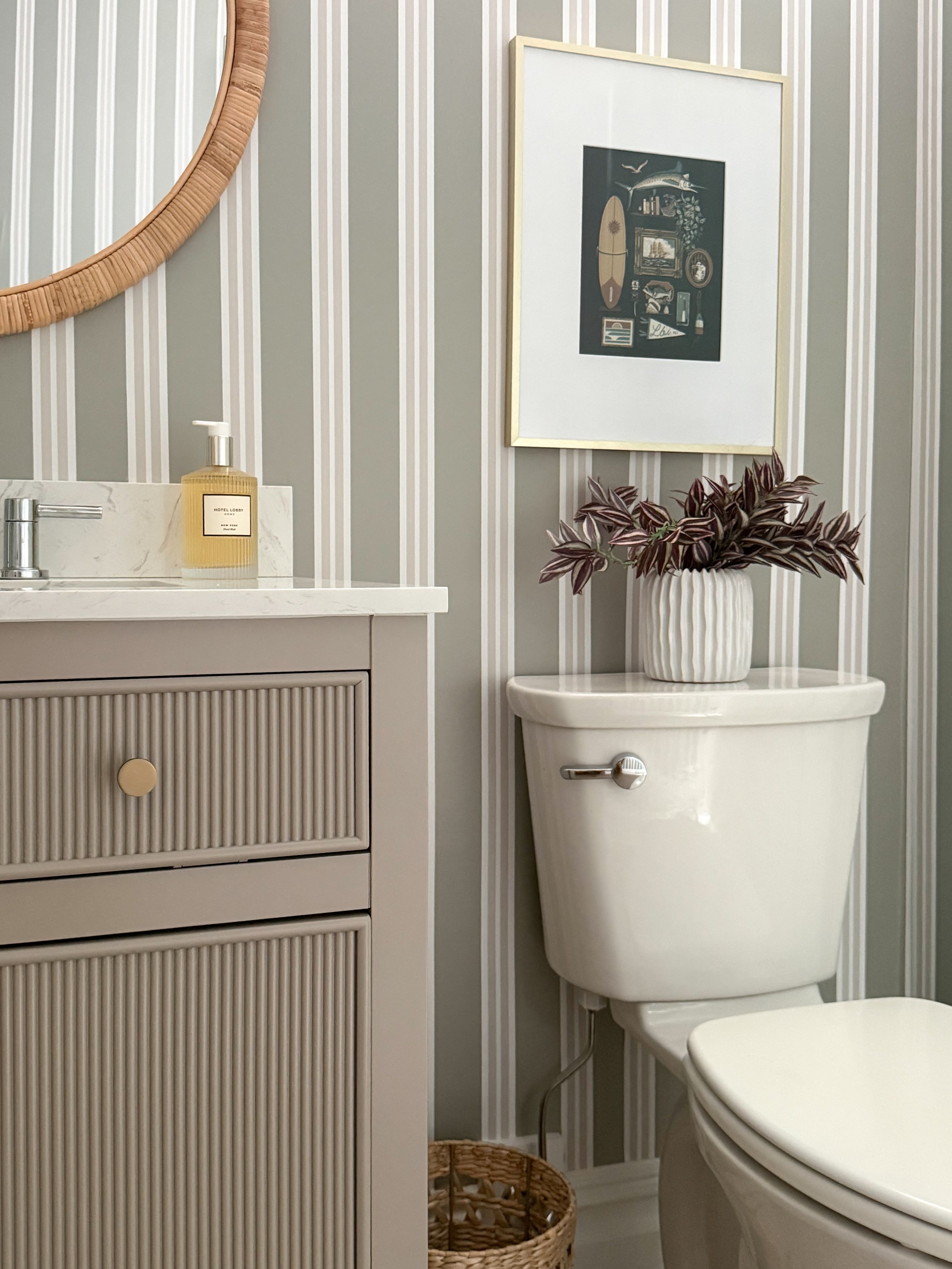 Small basement bathroom corner featuring Chasing Paper striped wallpaper, fluted vanity, framed artwork, decorative vase with greenery, and neutral bathroom styling.